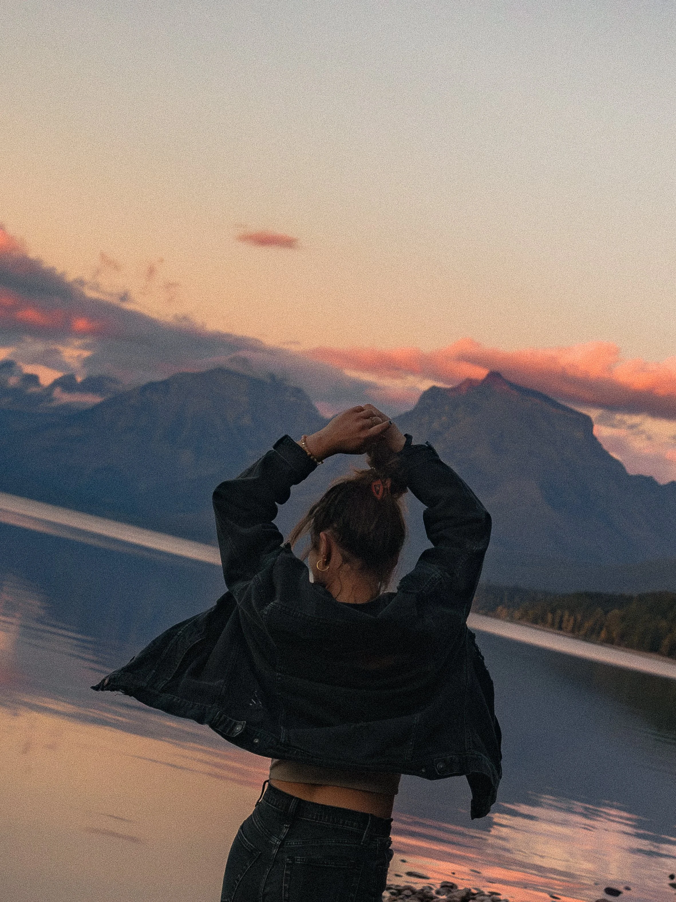 A woman standing by a lake at sunset with mountains in the background, wearing a black jacket and dark jeans, her hair tied up at Lake MacDonald in Glacier National Park, Montana