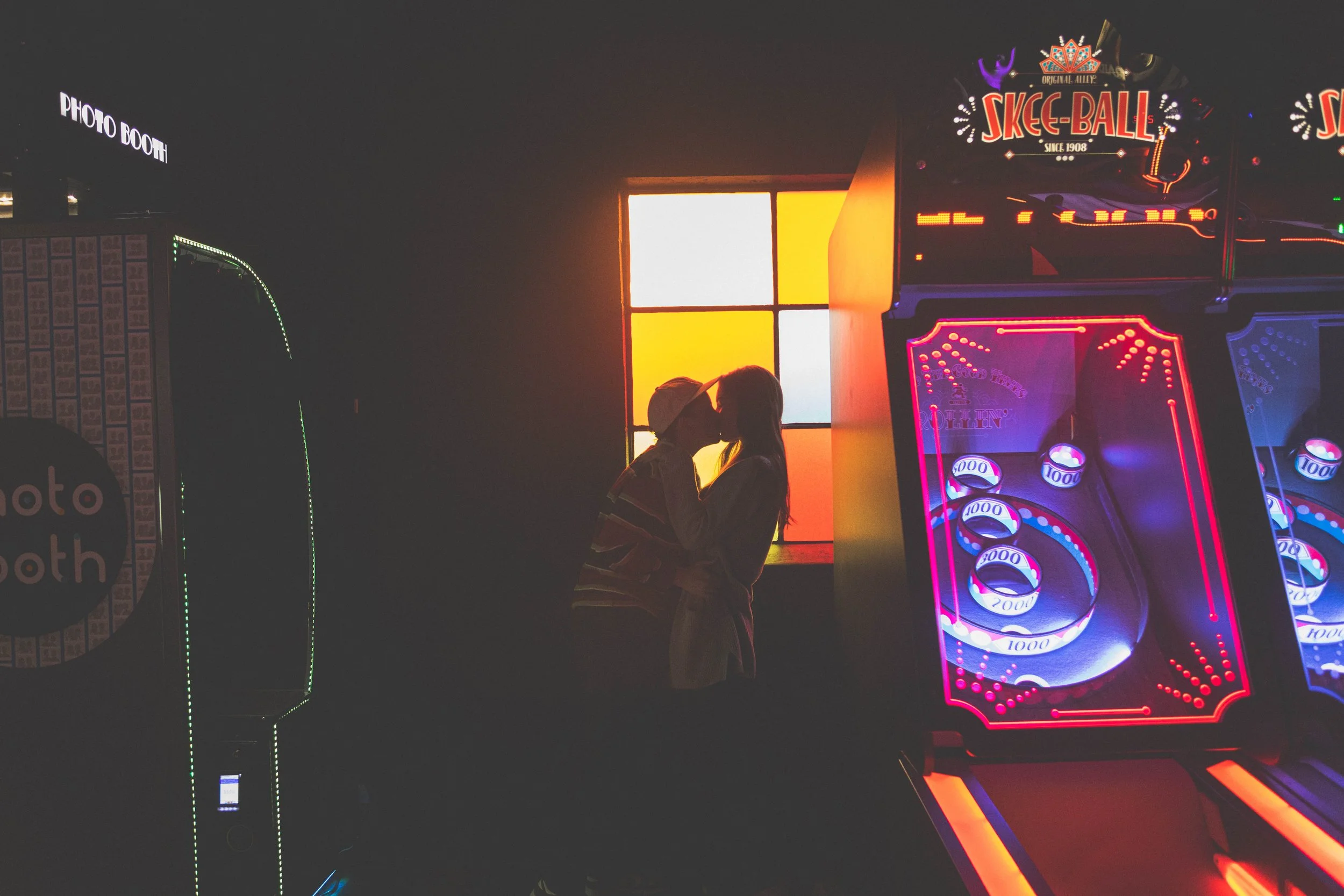 A silhouette of a couple kissing in front of a stained glass window with colorful panels at an arcade, with pinball machines and an arcade photo booth visible.