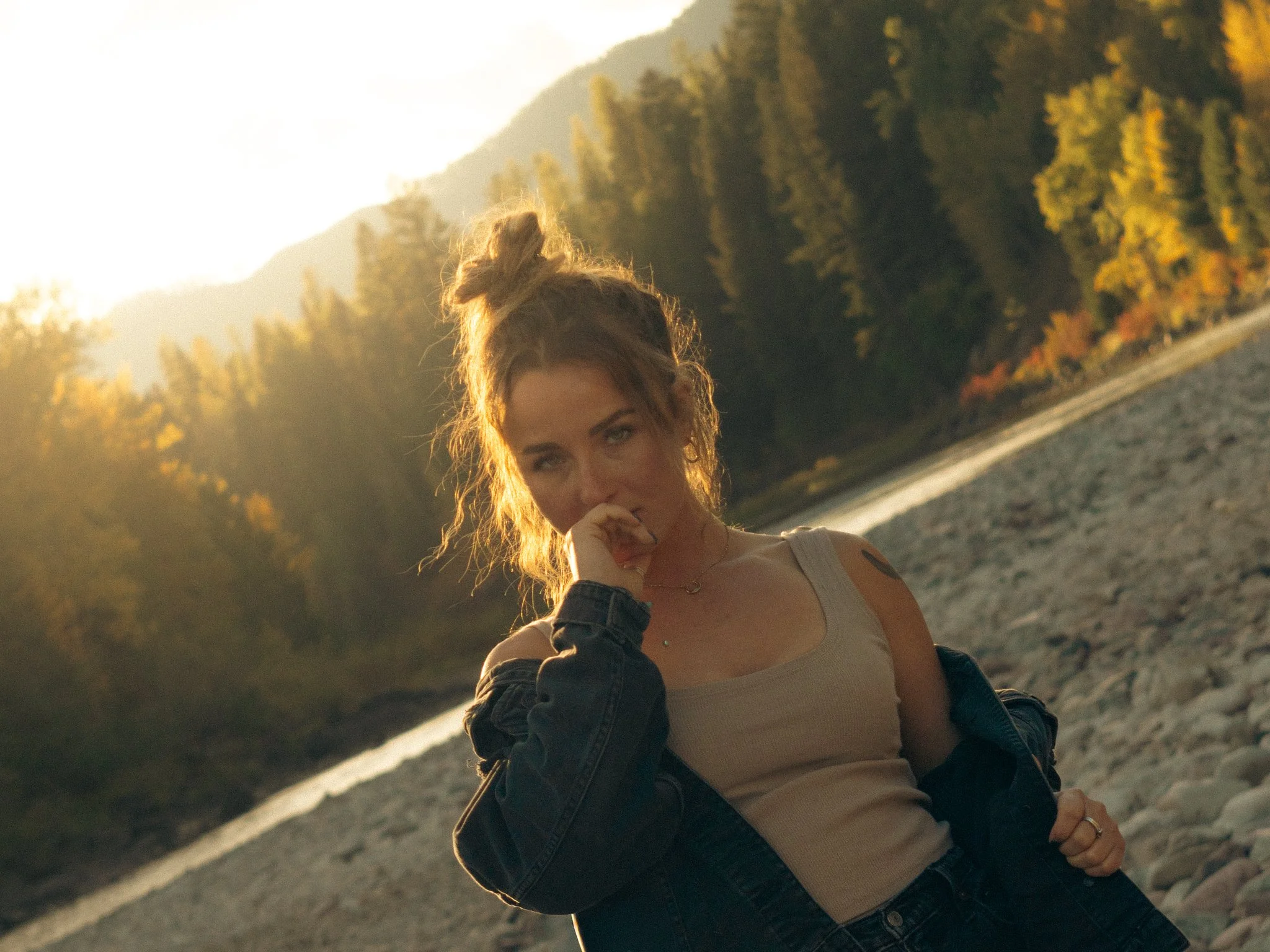 A woman with curly hair, wearing a beige tank top and black jacket, standing near a river at sunset with a forested mountain background.