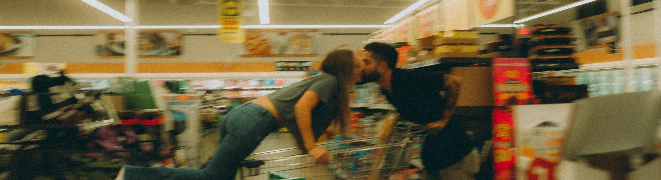 A young couple is sharing a kiss in a grocery store aisle next to a shopping cart.