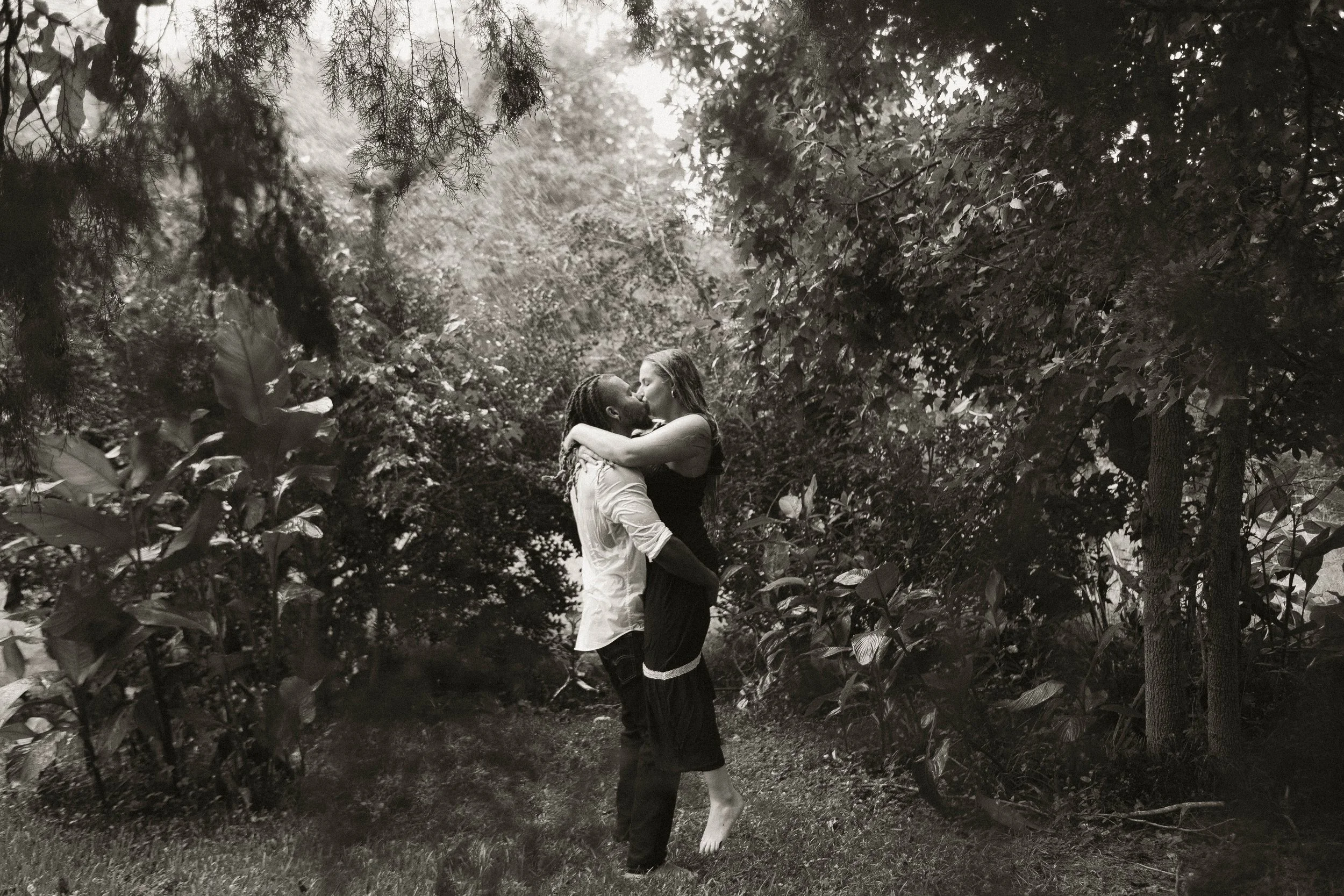 A couple embracing in a forested area, with the woman lifted off the ground by the man, surrounded by trees and foliage.