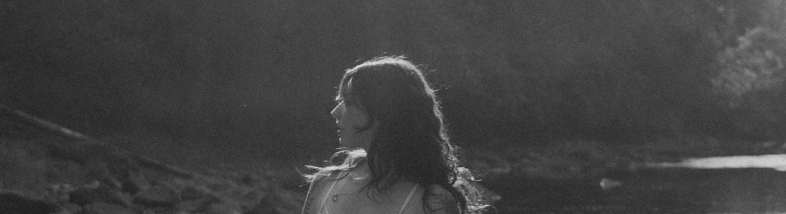 A woman with long, wavy hair looking to the side outdoors in sunlight, with a mountain or hillside background.