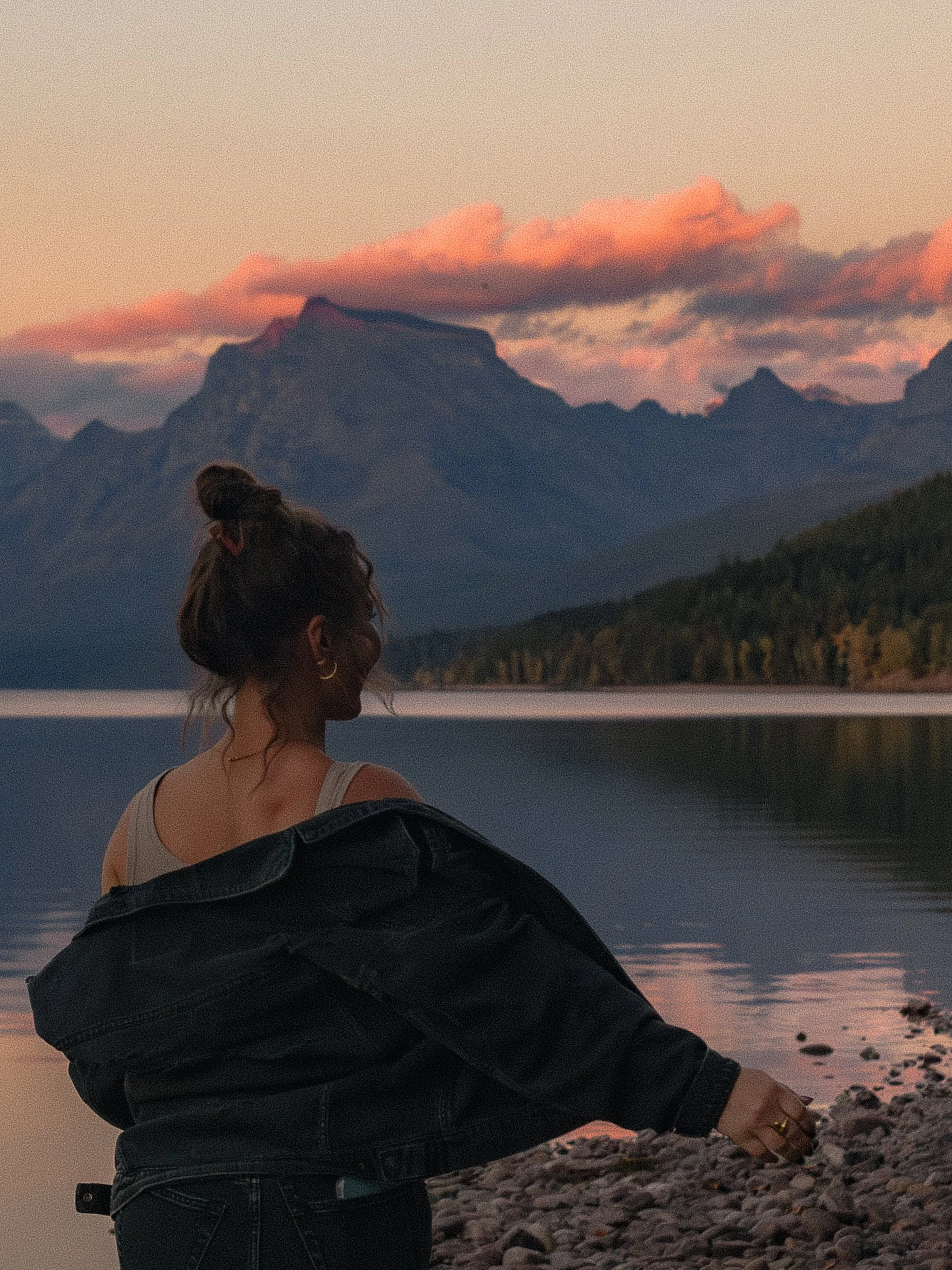 A woman sitting on a rocky shore by a lake during sunset, with mountains and pink clouds in the background.