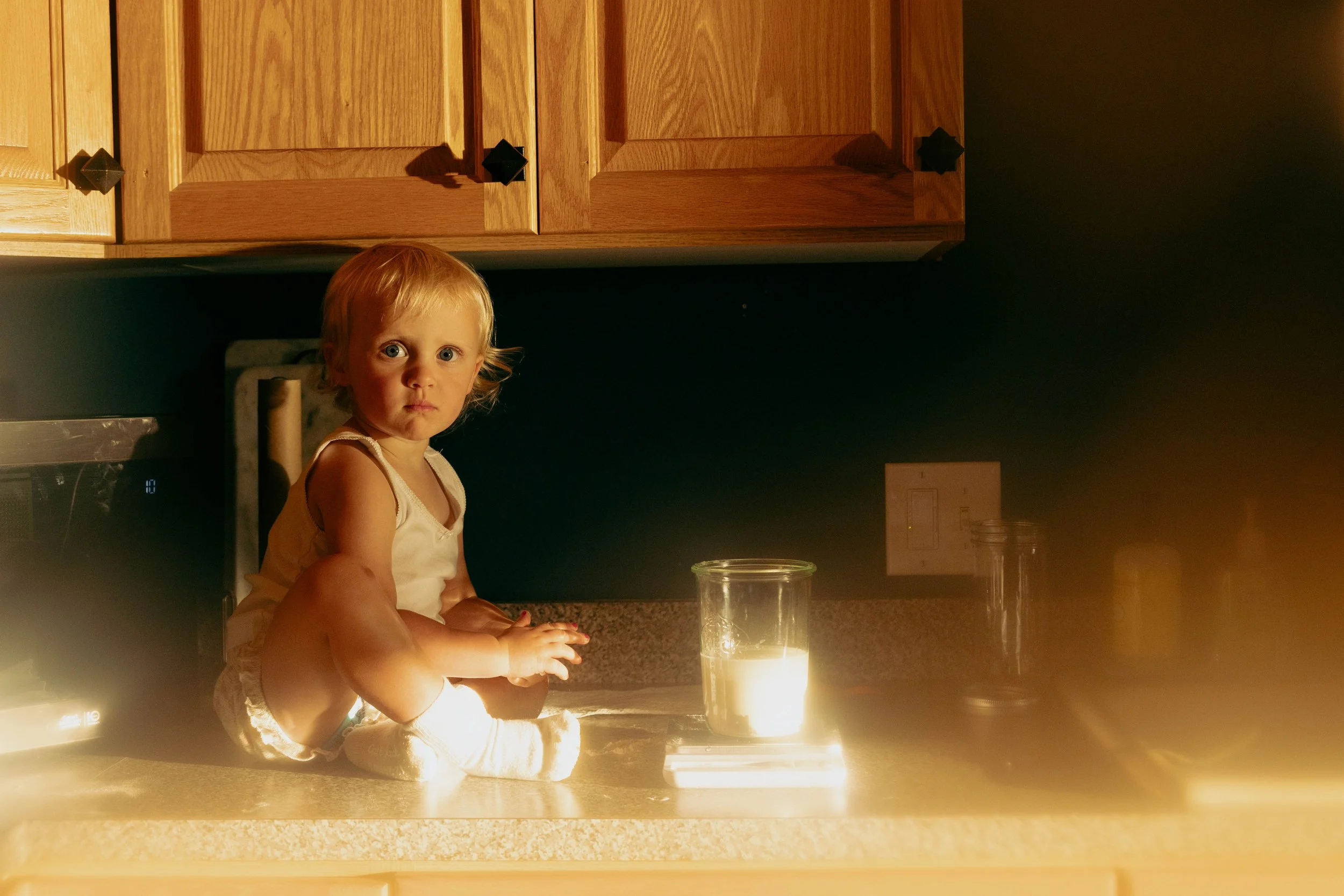 A young girl sitting on a kitchen counter illuminated by candlelight, looking at the camera with a serious expression.