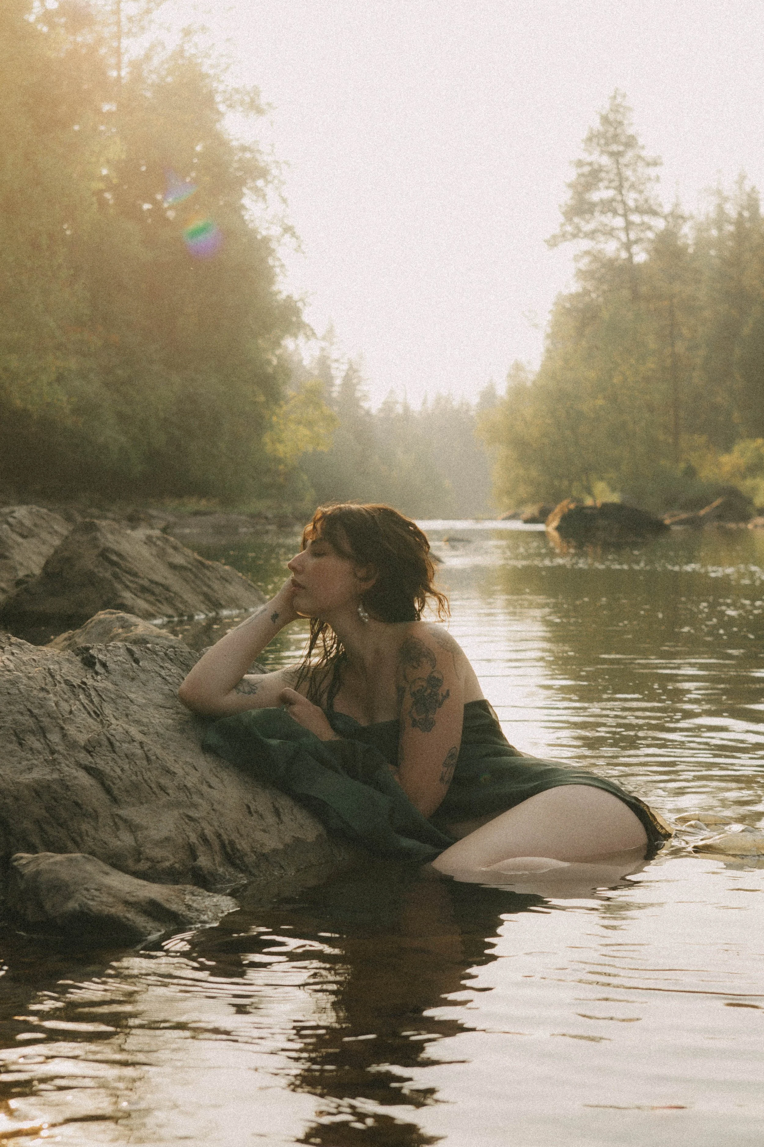 A woman with tattoos lying on a rock in a river, with trees and sunlight in the background.