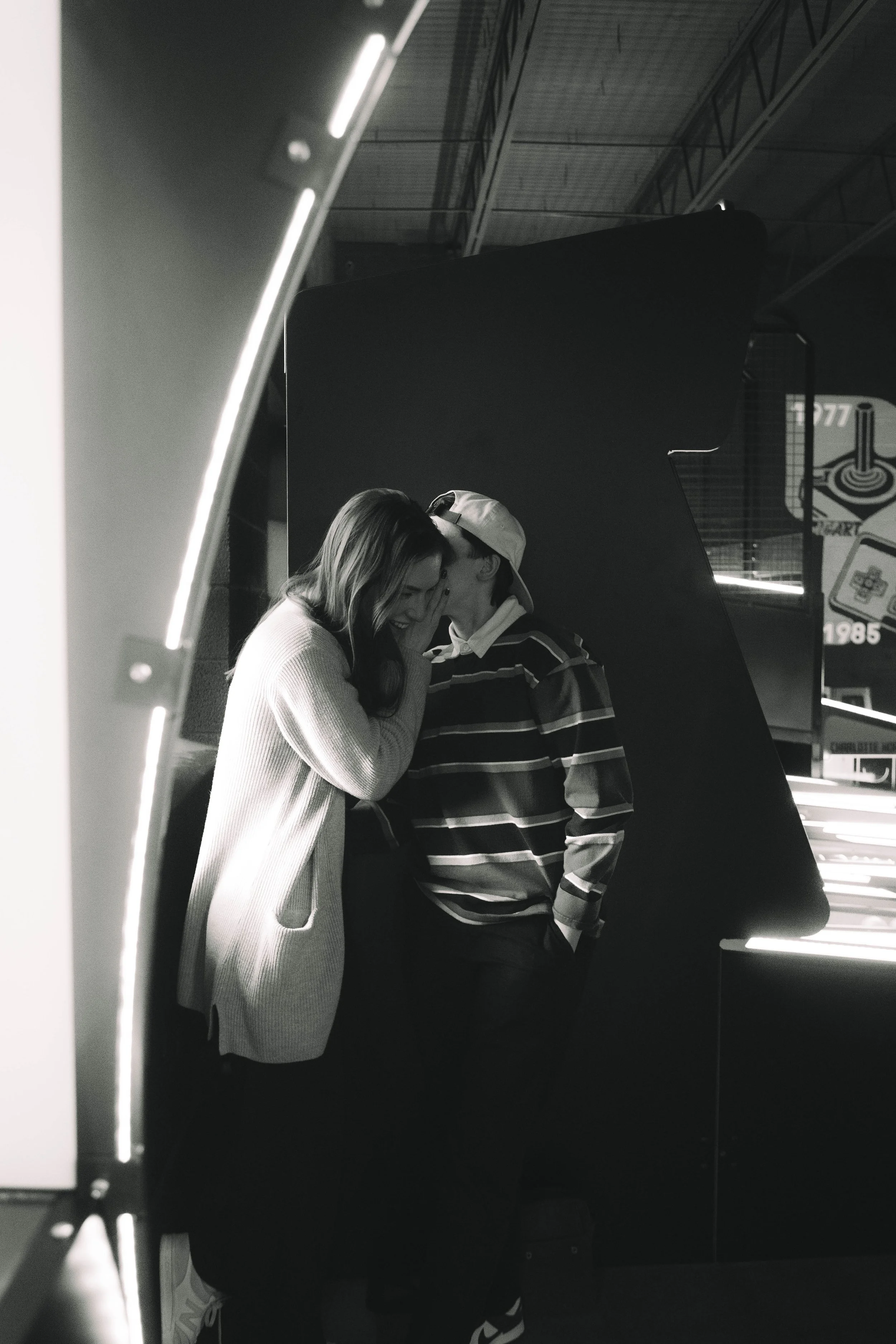 A black and white photo of a young couple sharing an intimate moment, smiling and leaning their foreheads together inside an arcade.