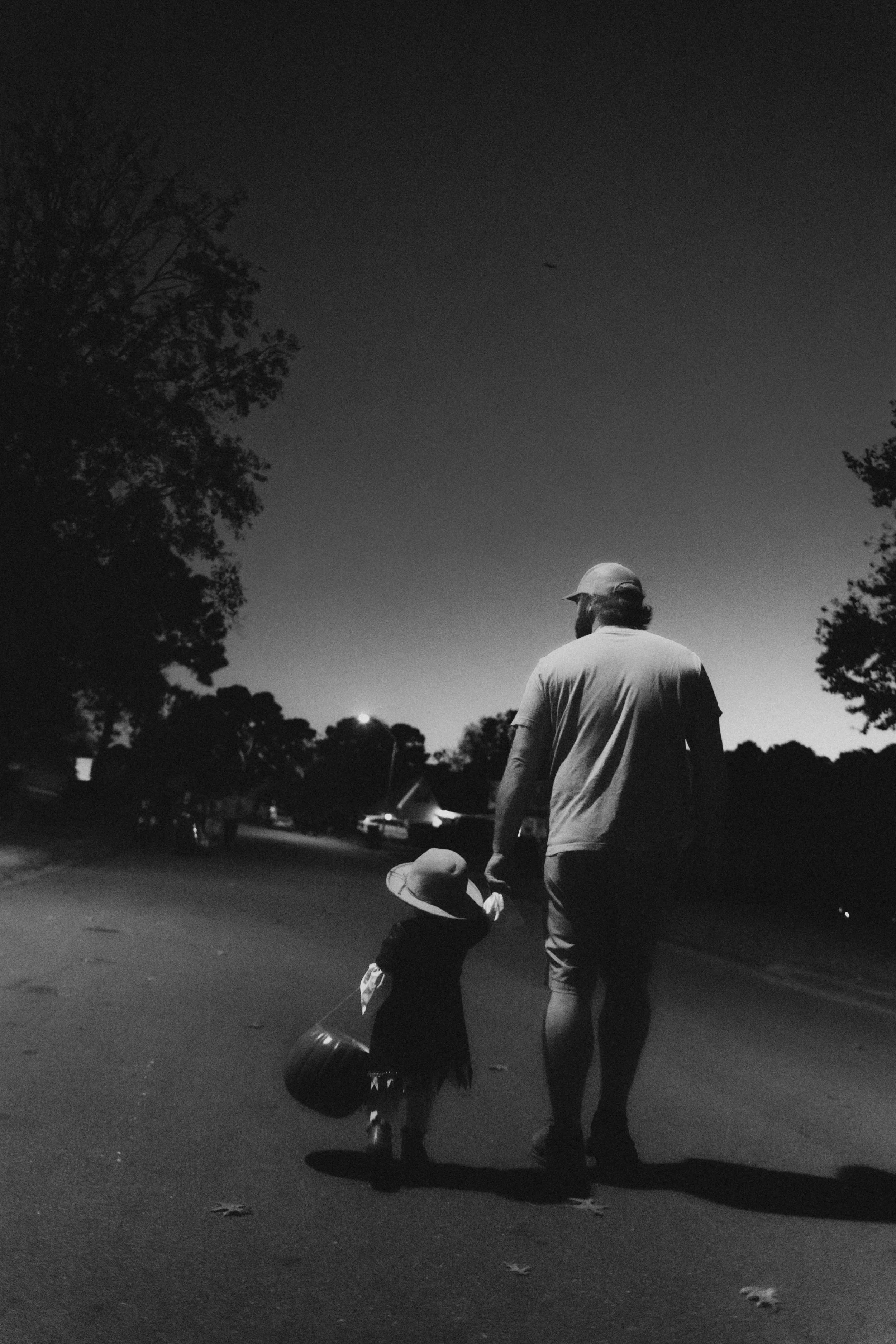 A man and a child walking on a suburban street at dusk, both wearing hats, with the man holding the child's hand.