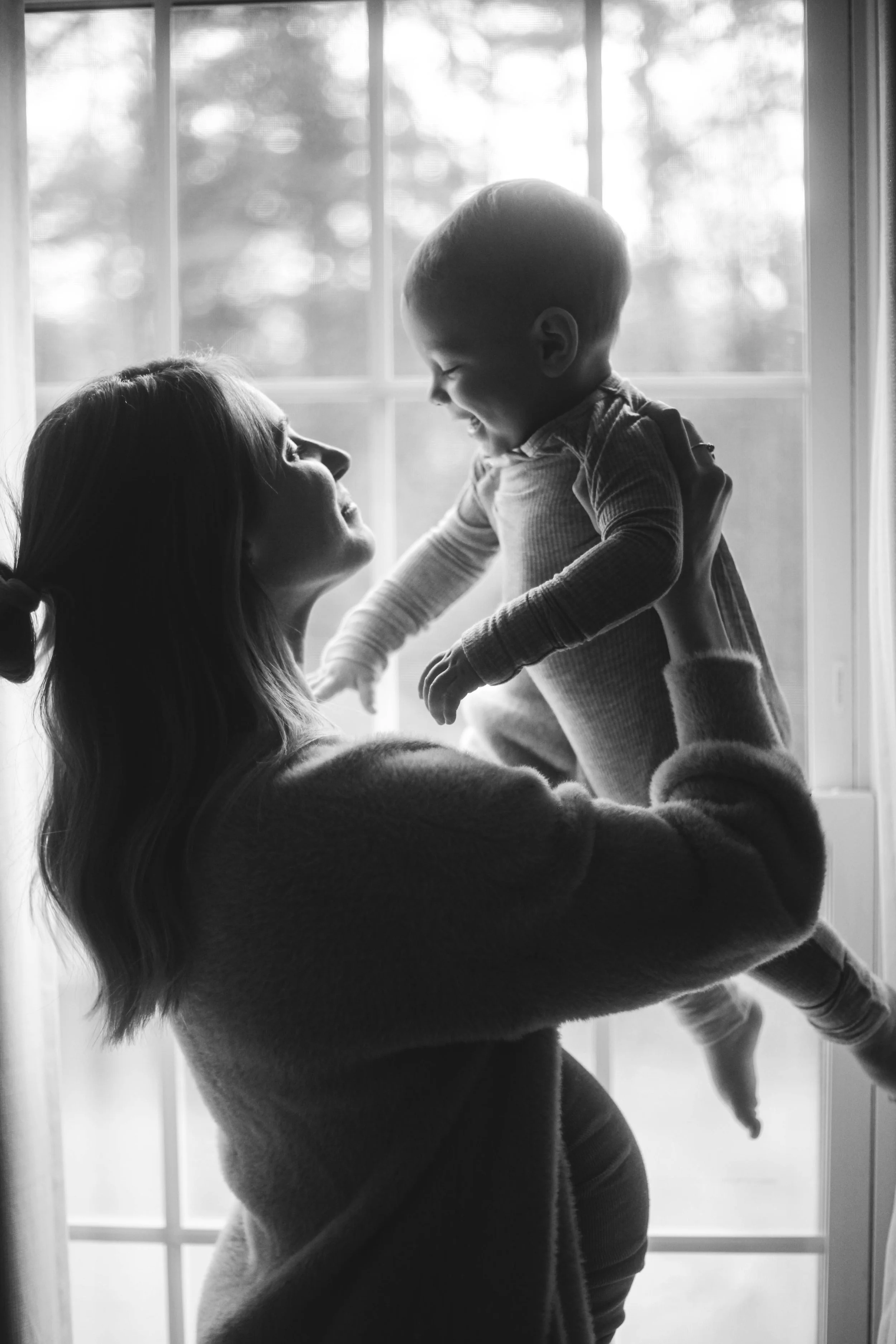 A woman lifting a smiling child in front of a bright window.