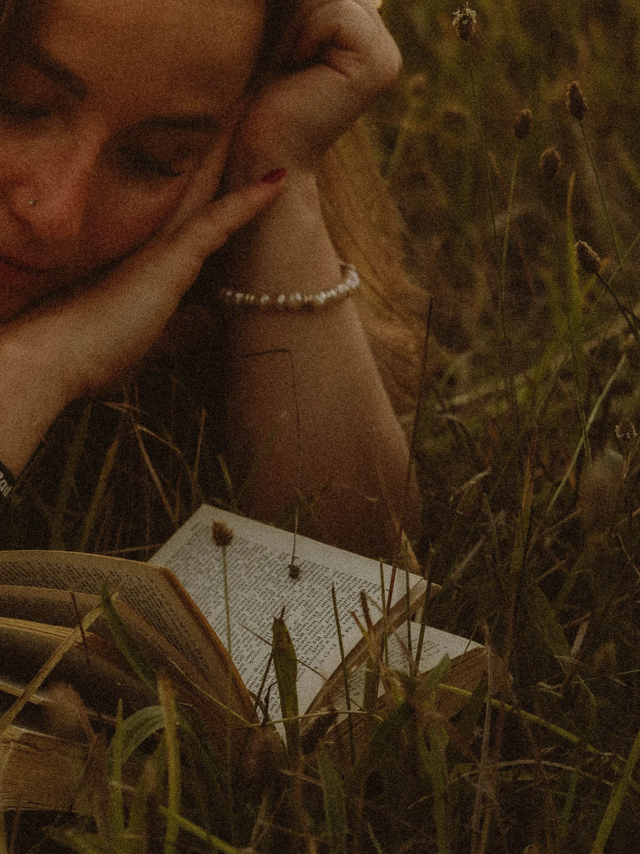A woman lying in grass with closed eyes, holding their head with one hand, wearing a pearl bracelet and a nose piercing, next to an open book during sunset.