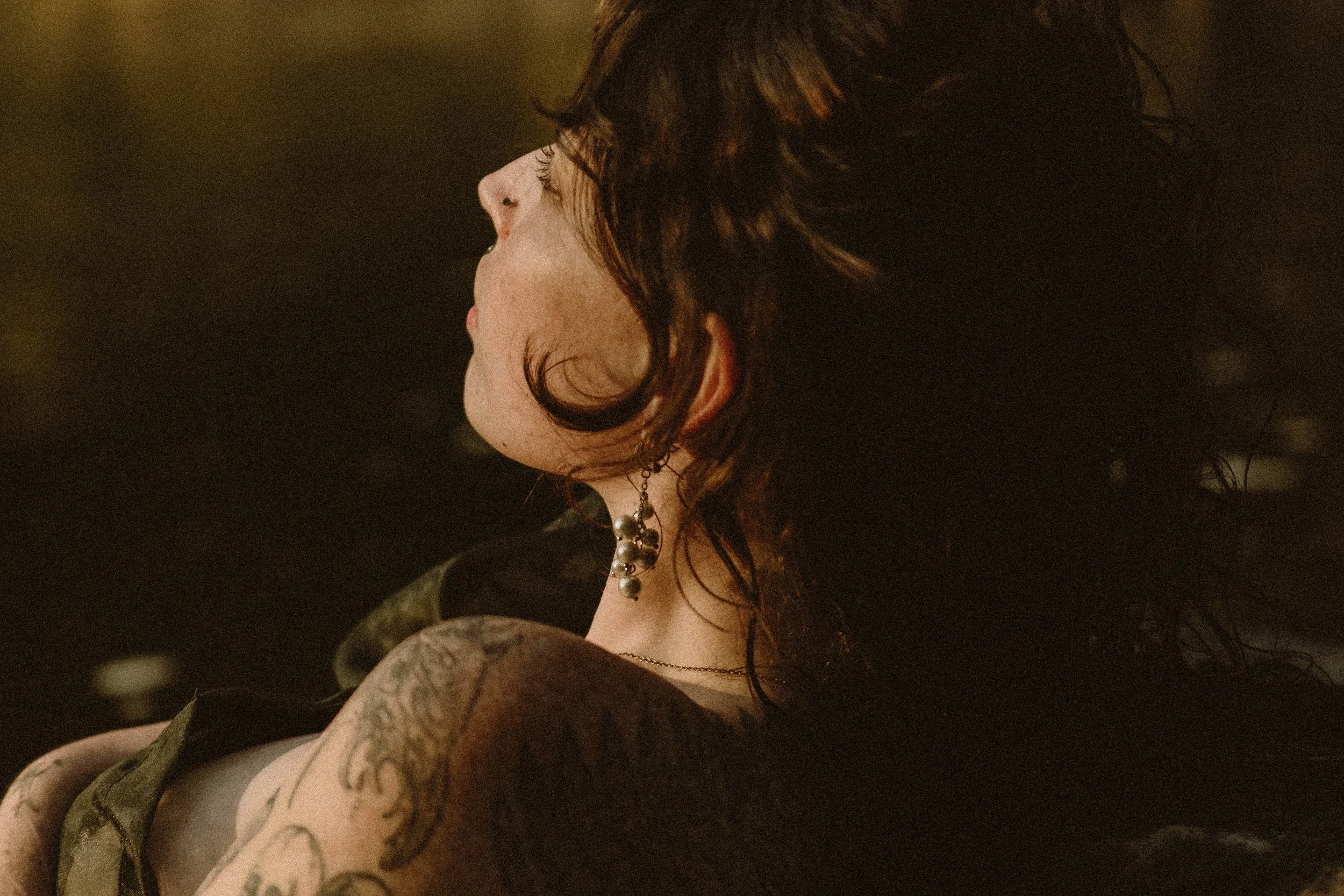 Close-up profile of a woman with dark, wavy hair, tattoos on her arm, wearing a necklace with large beads, in a dimly lit river.