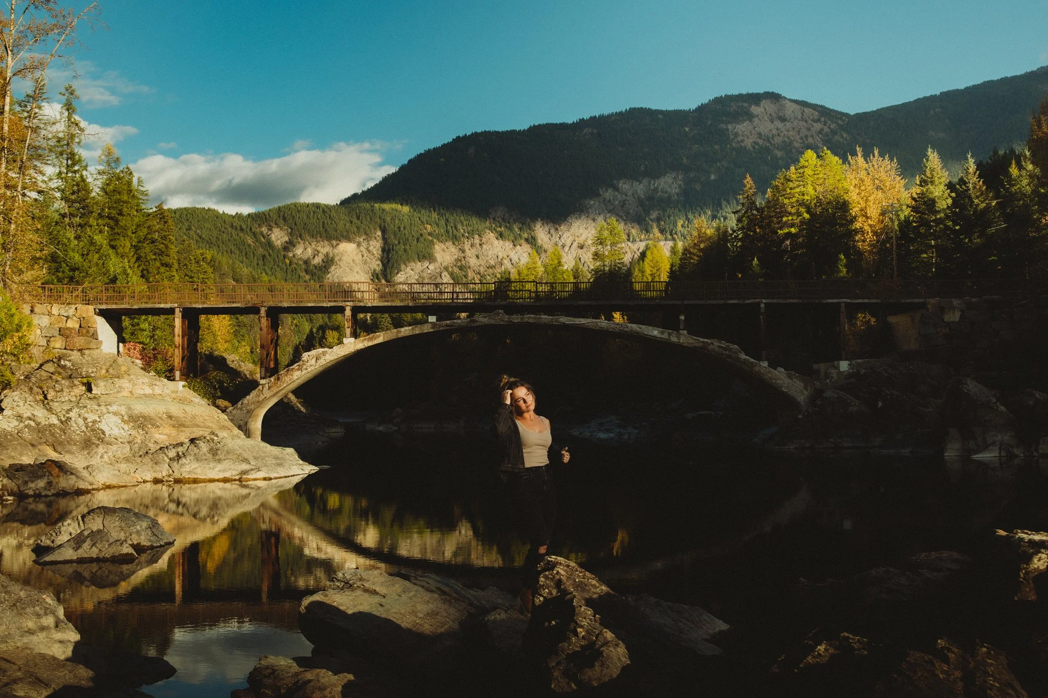 A woman standing on rocks in a river with an arched stone bridge and forested mountains in the background at Glacier National Park during daylight.