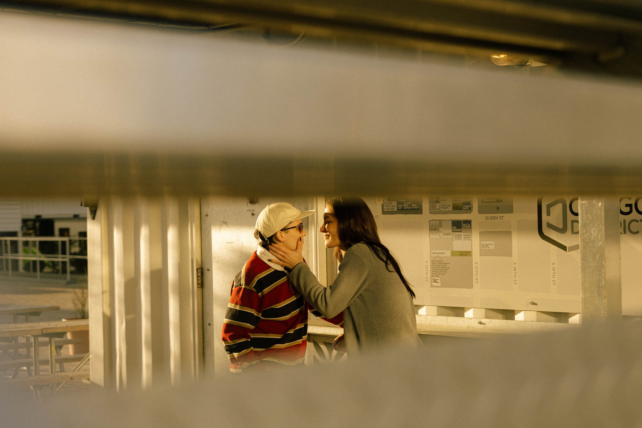Two women are engaged in an intimate moment, while leaning close, outside at a bar. Framed between horizontal metal bars.