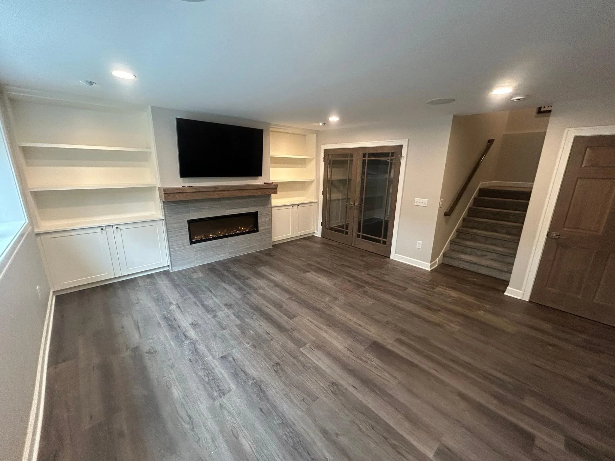 Empty living room with wood flooring, built-in white shelves, a flat-screen TV, a fireplace with a wooden mantle, glass double doors, and staircase with wooden steps.
