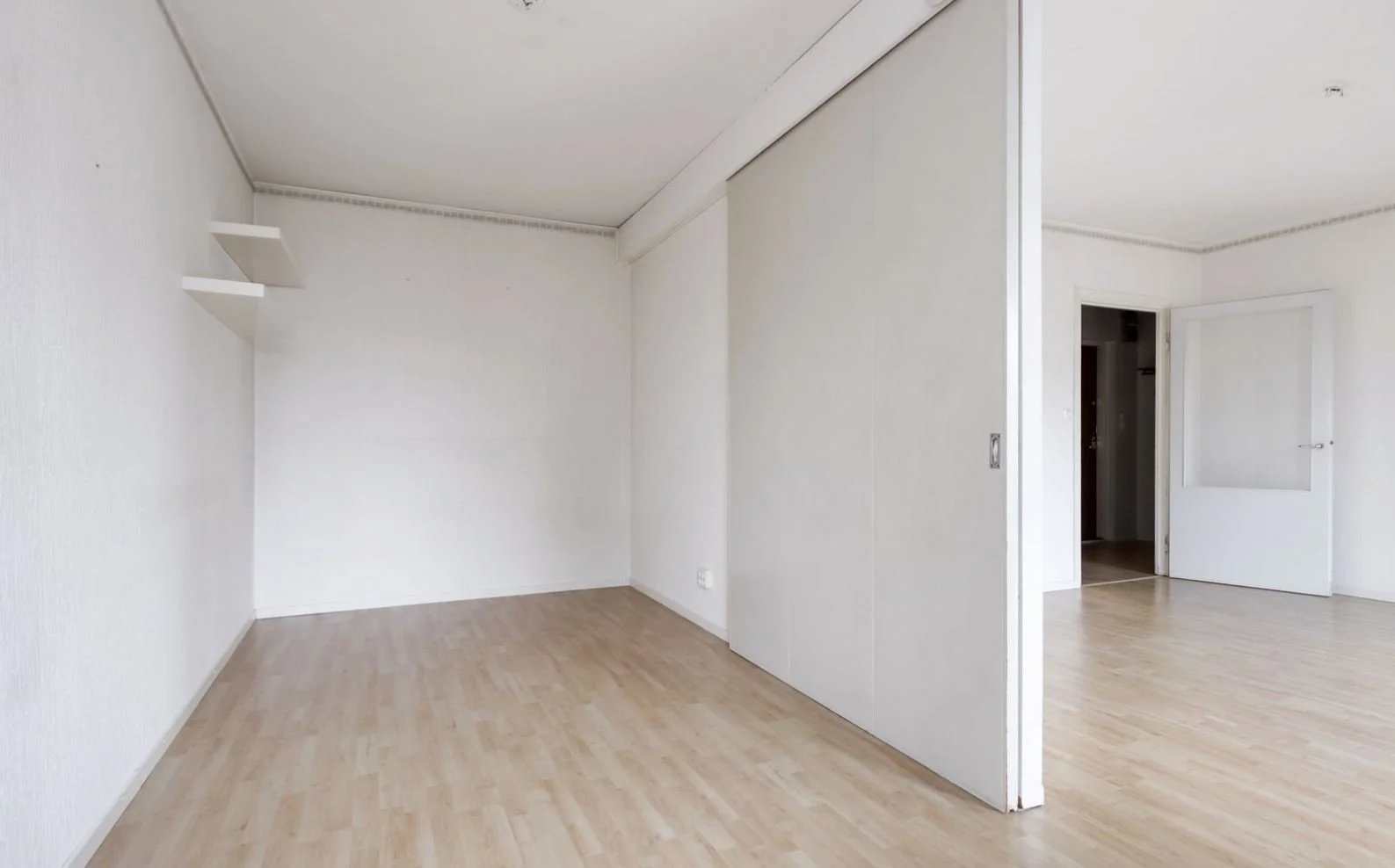 Pièce vide avec murs blancs, sol en bois clair, porte ouverte vers une autre pièce, quelques étagères blanches sur le mur gauche.