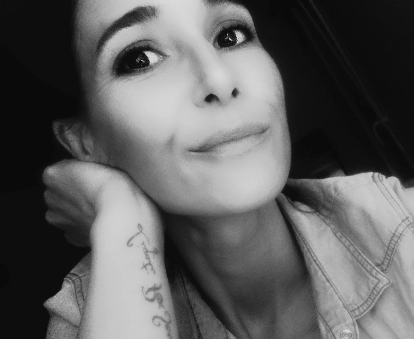 Portrait en noir et blanc d'une femme souriante, avec une main derrière la tête, portant une veste en jean, tatouage sur l'avant-bras.