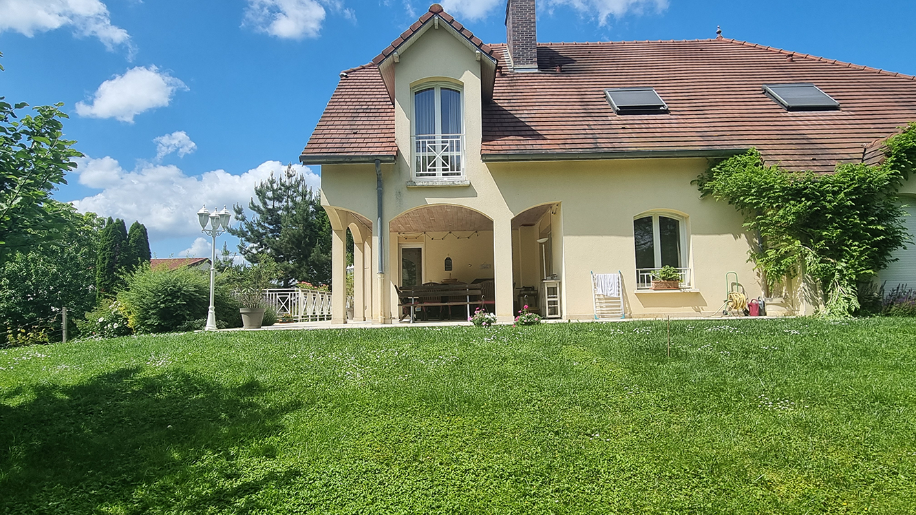 Maison individuelle beige avec toiture en tuiles rouges, balcon au premier étage, terrasse arrière, jardin avec pelouse, arbres et mobilier extérieur sous un ciel bleu avec quelques nuages.