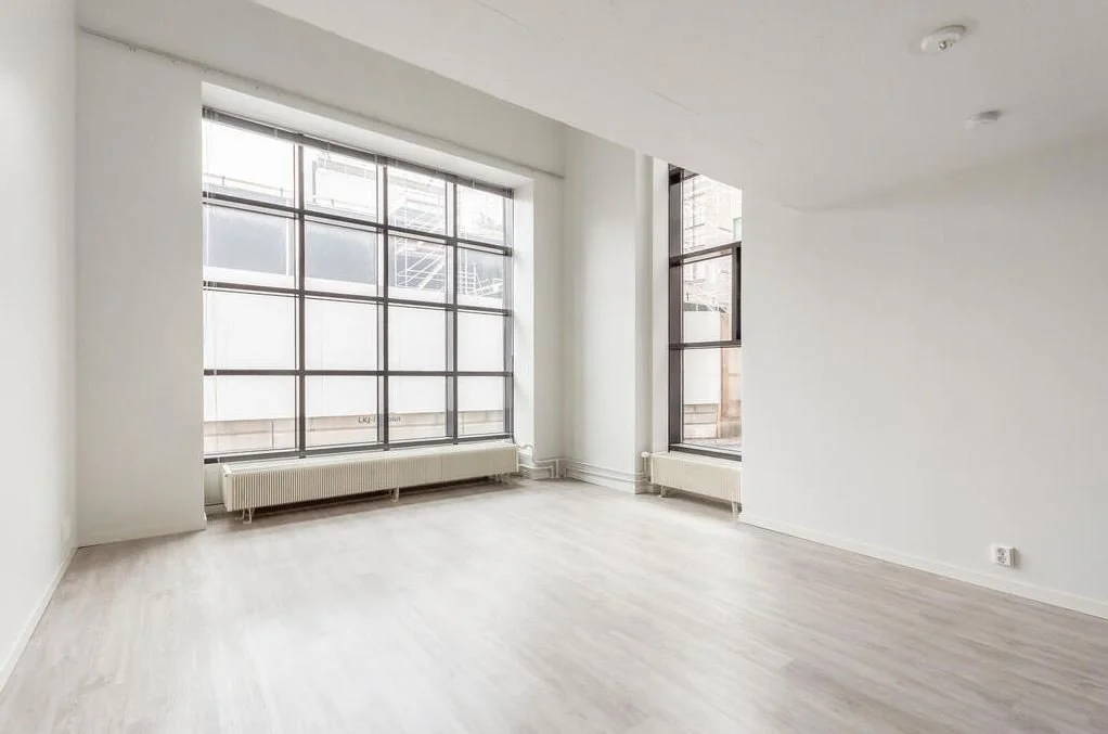 Une pièce vide avec de grandes fenêtres laissant entrer beaucoup de lumière, murs blancs et sol en bois clair.