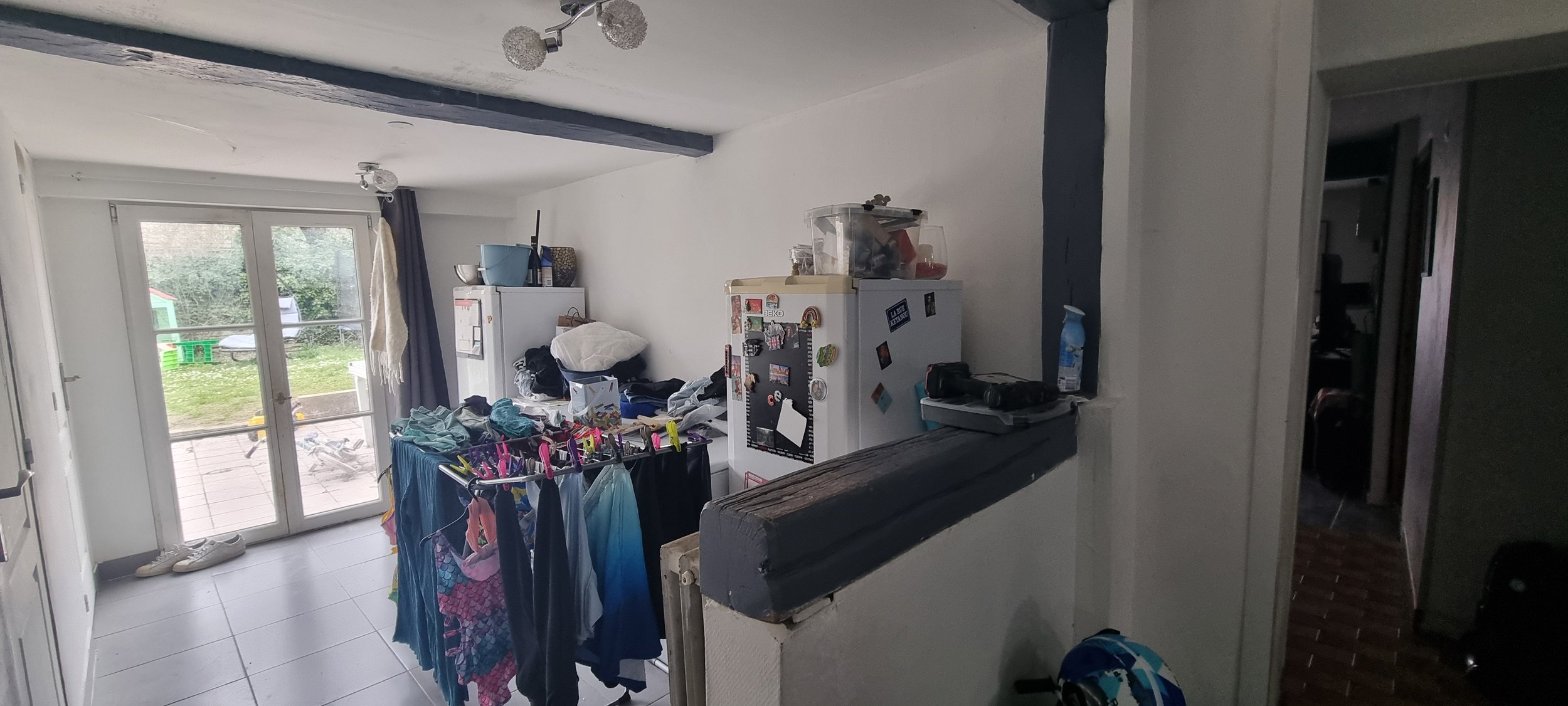 Pièce d'habitation avec une grande fenêtre donnant sur un jardin, une table avec du linge étendu, des meubles et des objets divers