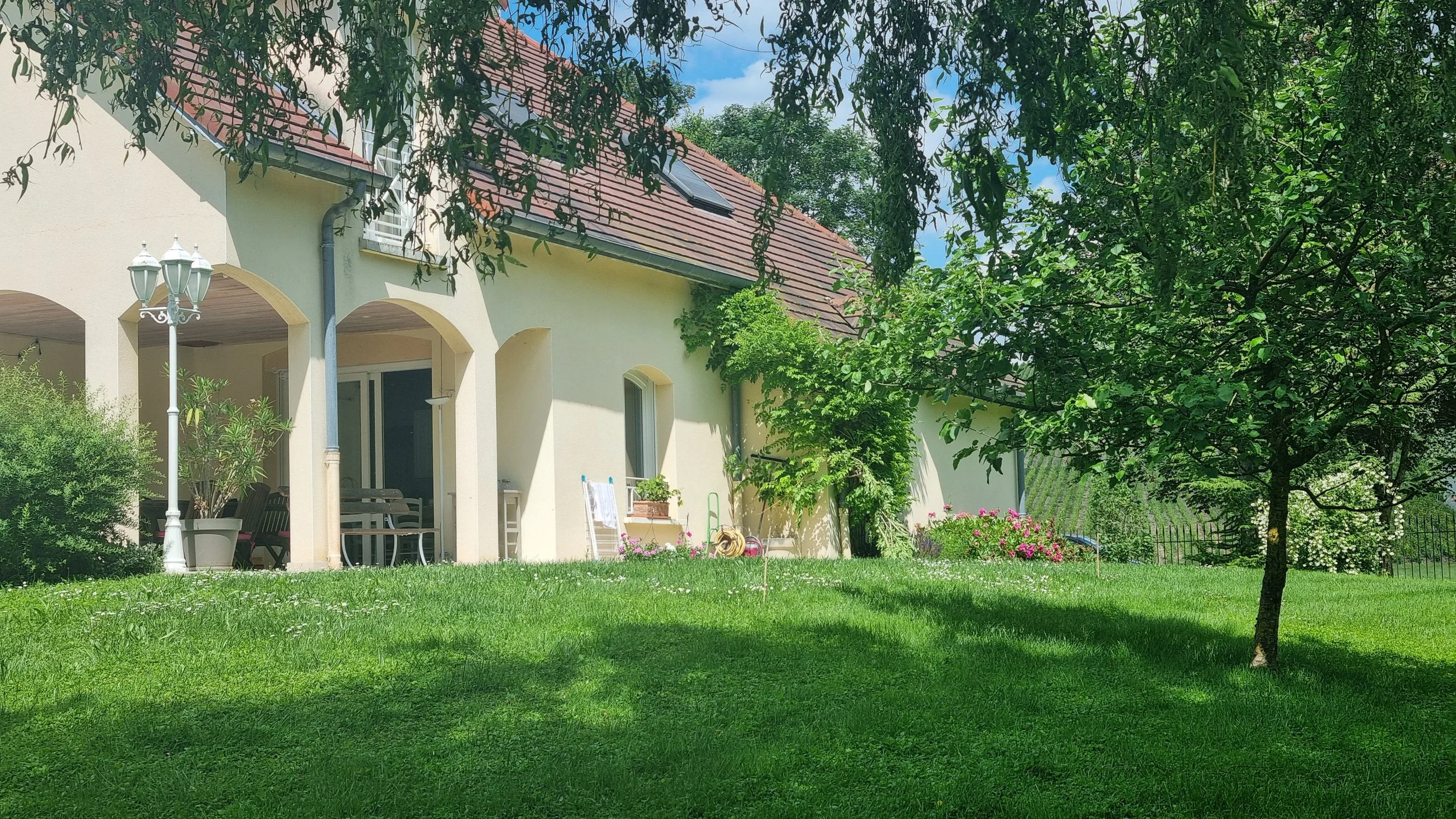 Une maison avec un toit en tuiles rouges, entourée d'un jardin vert avec des arbres et des fleurs, sous un ciel bleu avec quelques nuages.