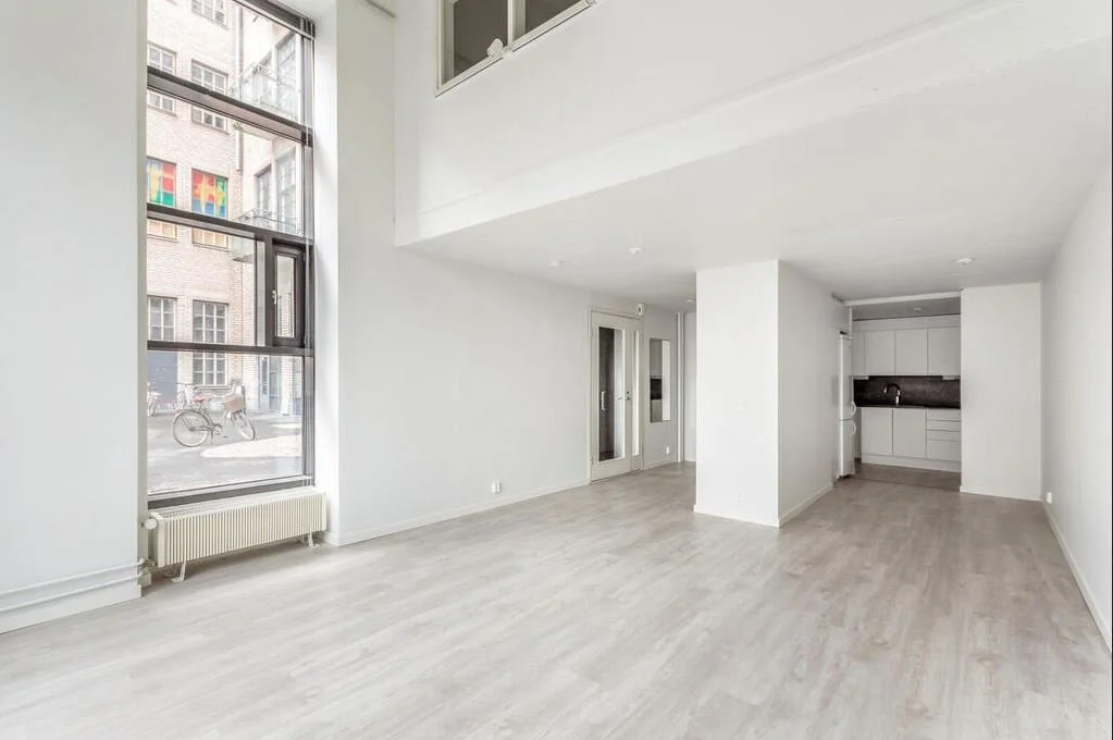 Un intérieur d'appartement avec de grandes fenêtres laissant entrer beaucoup de lumière naturelle, murs blancs, sol en bois clair, cuisine compacte en arrière-plan.