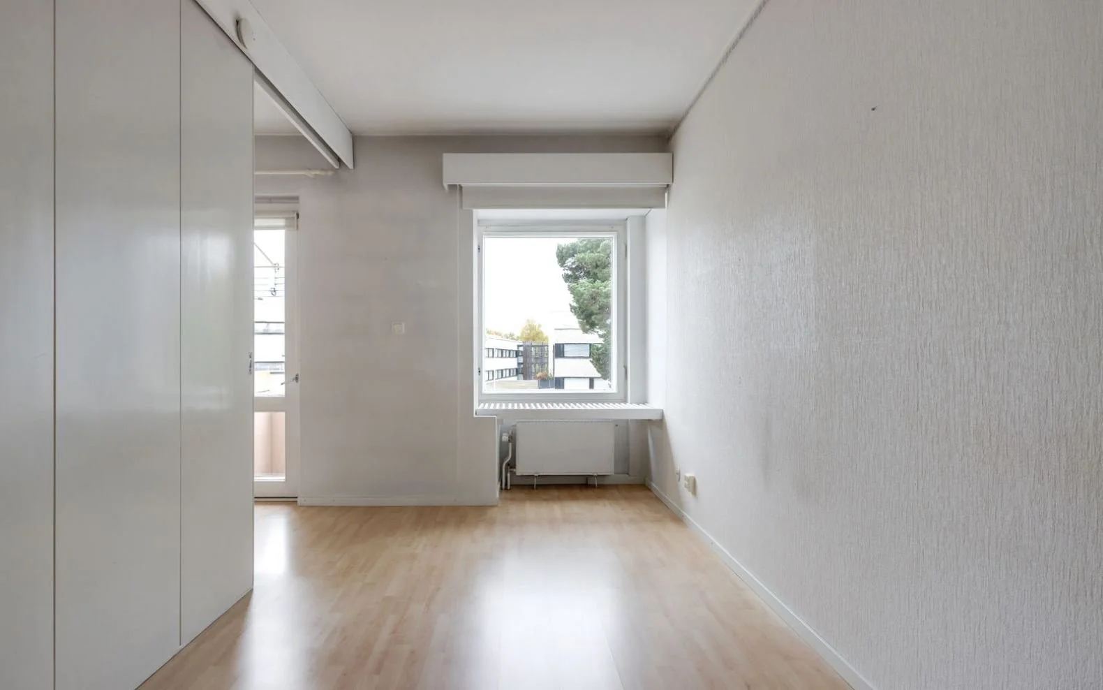 Pièce vide avec parquet en bois clair, fenêtre donnant sur l'extérieur avec un arbre, placard blanc intégré, radiateur sous la fenêtre, mur blanc.