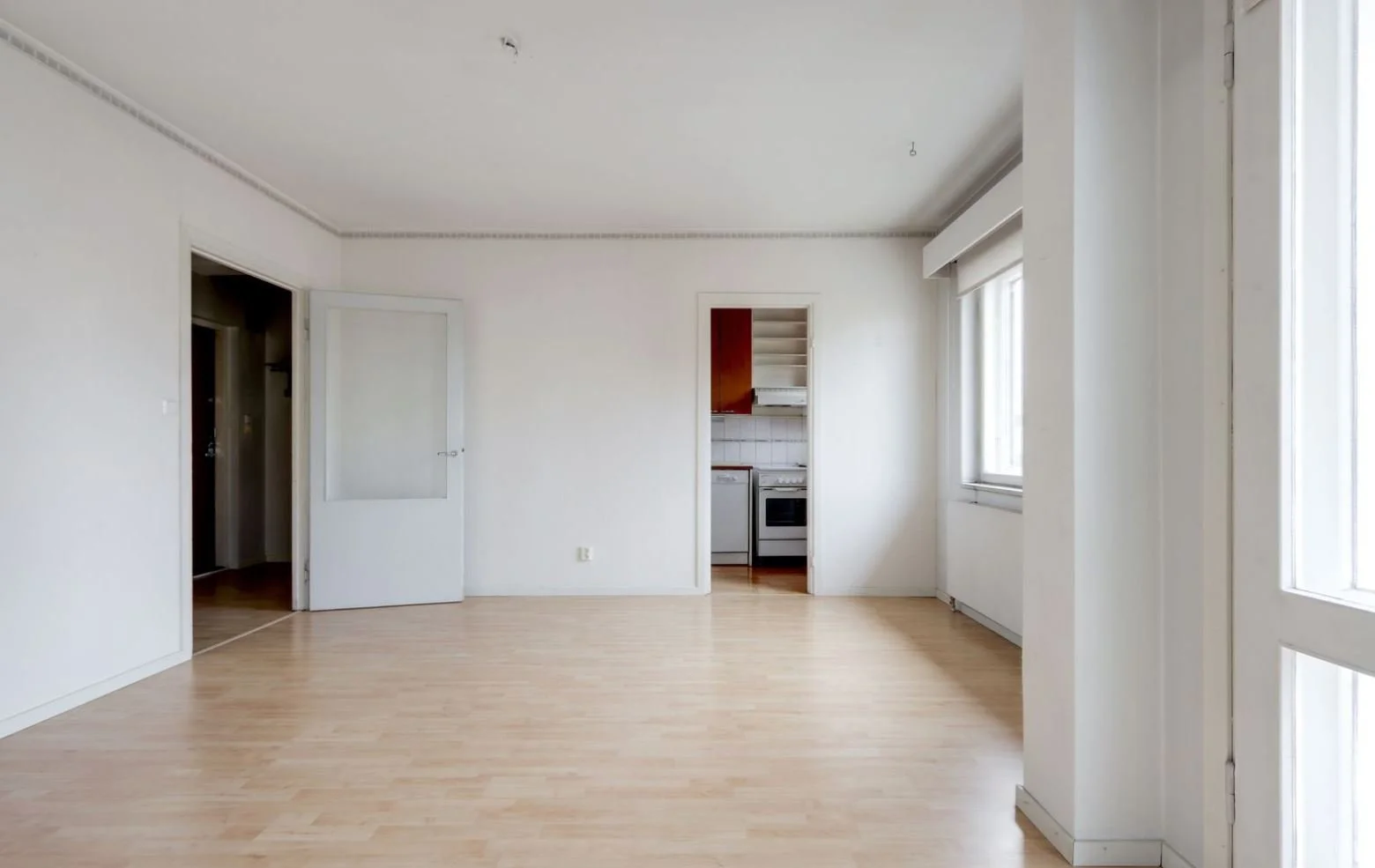 Appartement vide avec murs blancs, sol en bois clair, cuisine ouverte avec four et frigo, fenêtres laissant entrer la lumière naturelle.