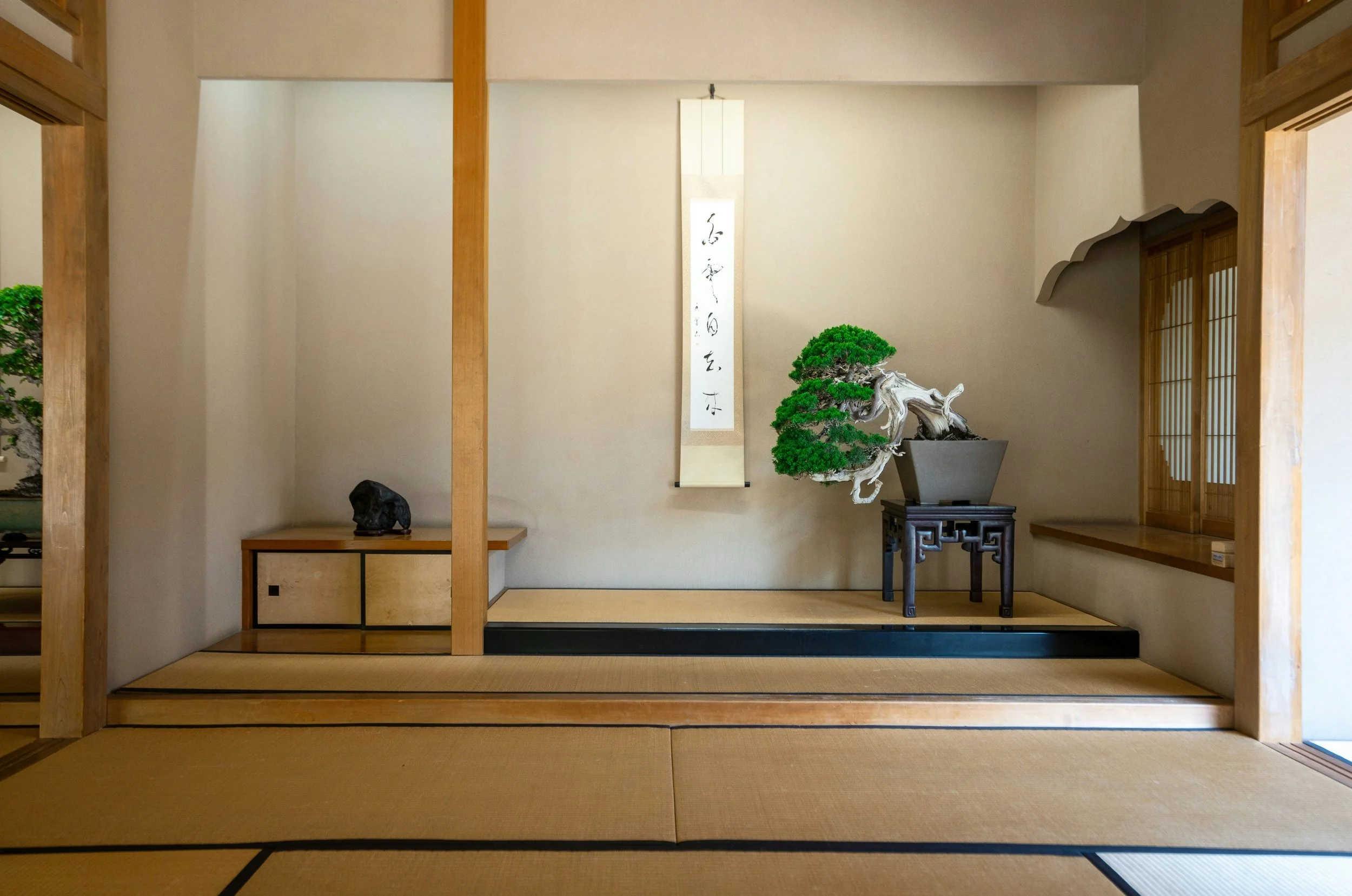 Intérieur d'une pièce de style japonais avec tatamis, un bonsaï sur un support en bois, une calligraphie suspendue sur le mur, et des éléments en bois traditionnels.