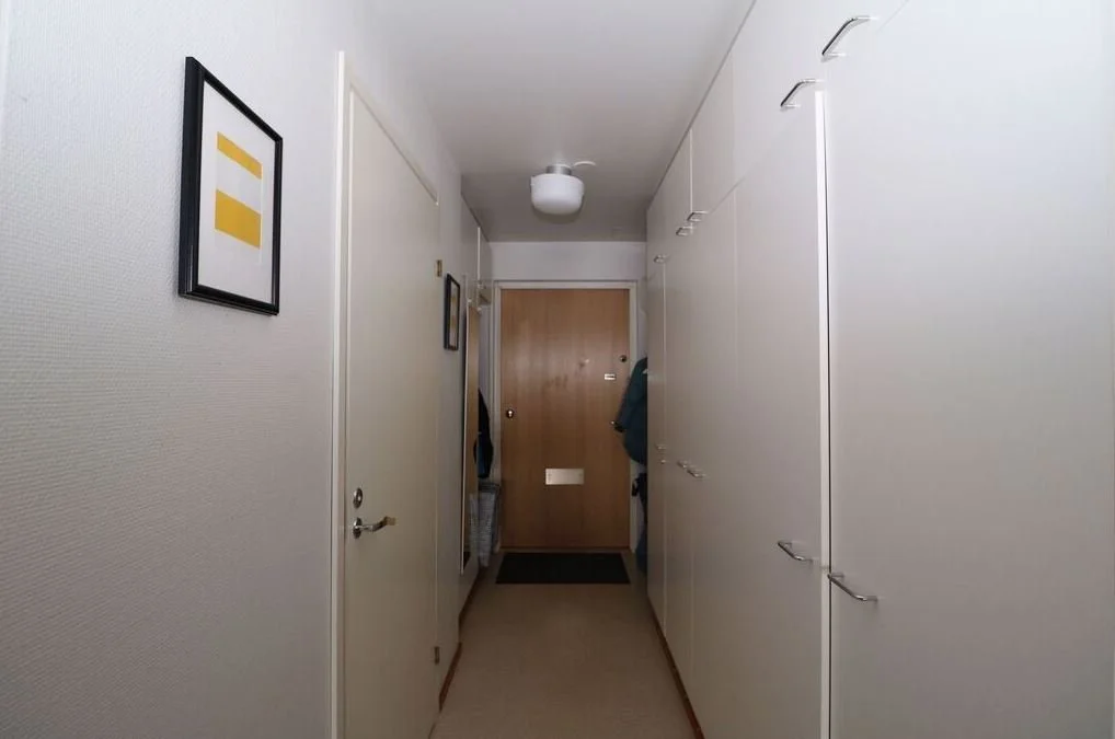 Vue d'un couloir intérieur avec des murs blancs, un placard à gauche, des cadres colorés accrochés au mur, un porte-manteau avec des vêtements, et une porte d'entrée en bois en fond.