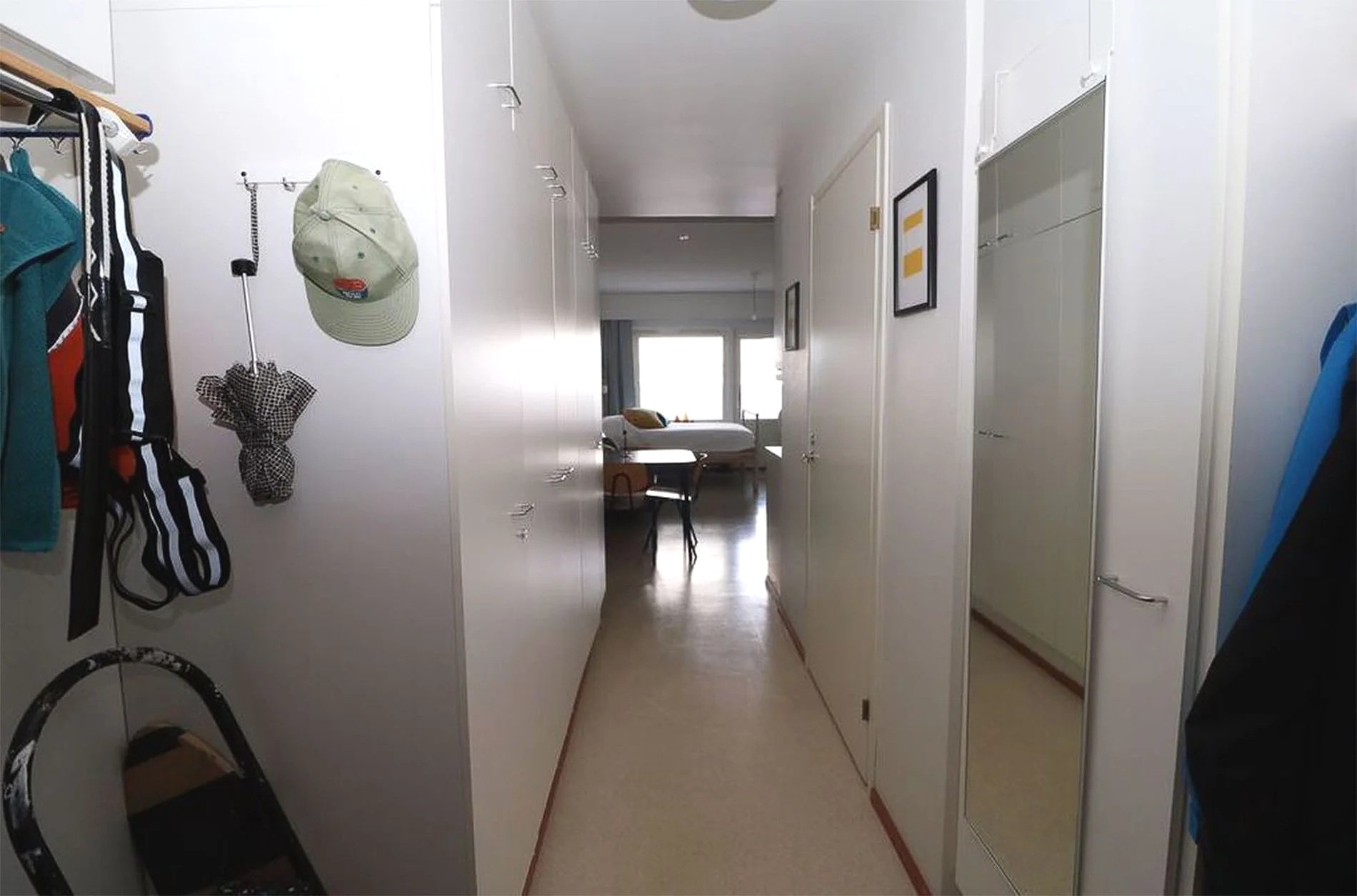 Couloir d'un appartement avec des murs blancs, un lit, une fenêtre, une table et une chaise au fond.