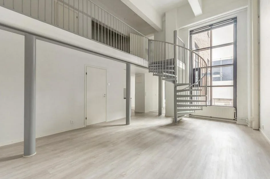 Intérieur lumineux avec une grande fenêtre, un escalier en colimaçon en métal, poutre apparente en haut, sol en bois clair.