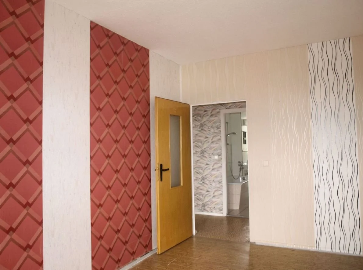 Intérieur d'une pièce avec des murs décorés de différents papiers peints, une porte en bois jaune menant à une salle de bain avec une baignoire et une douche.