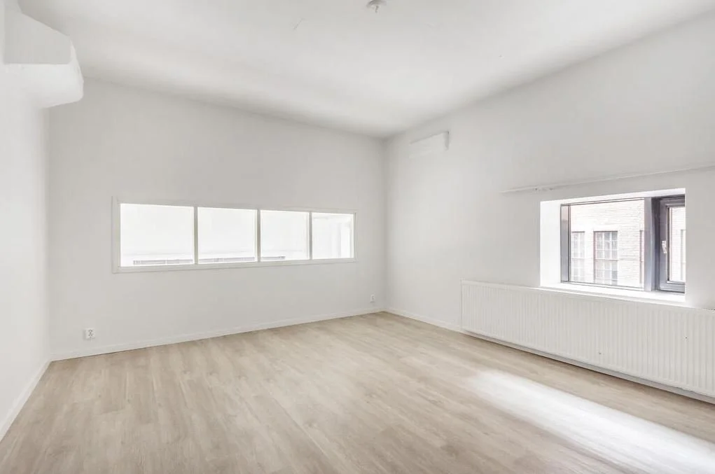 Une pièce vide avec des murs blancs, deux fenêtres, un radiateur et un sol en bois clair.