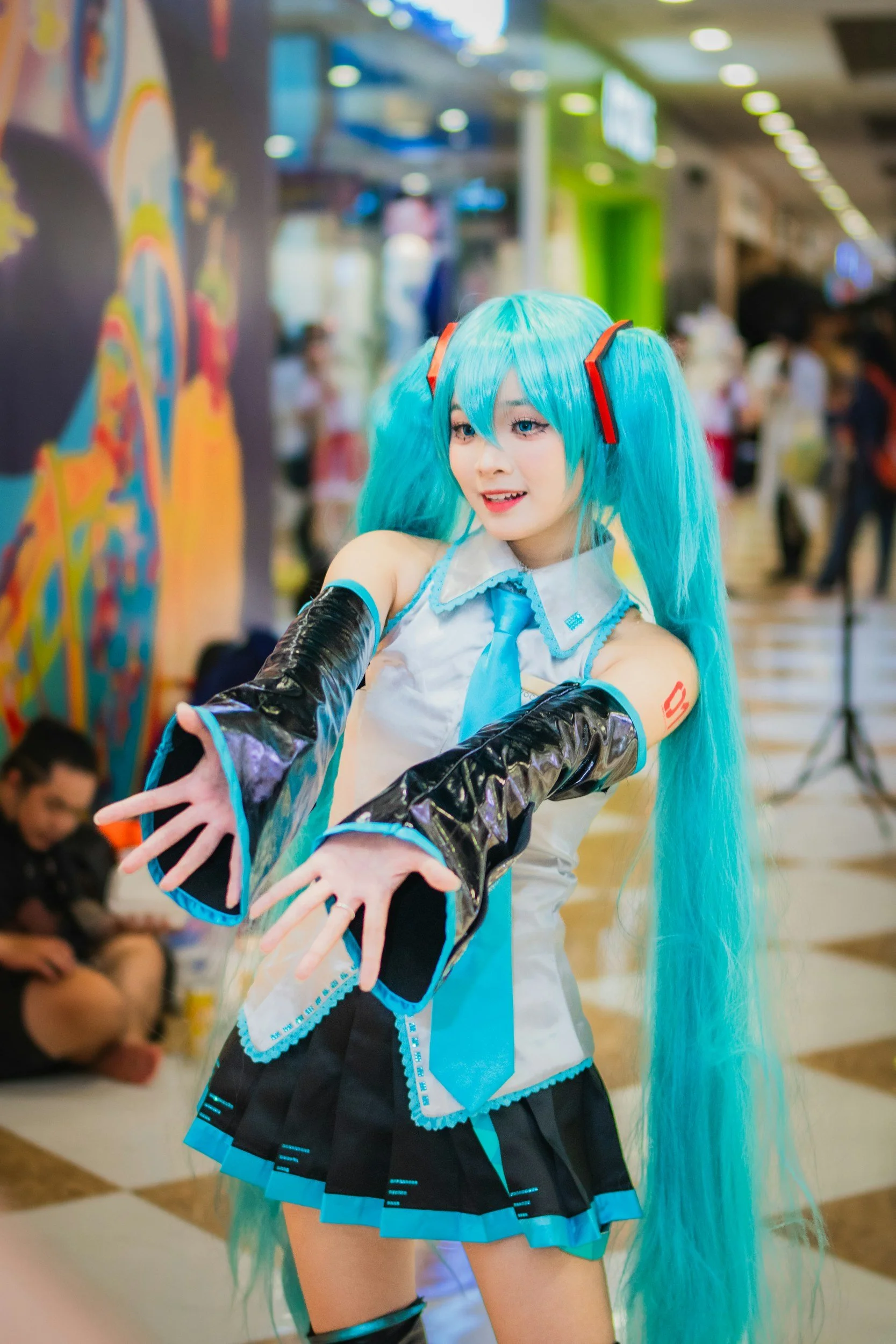 A cosplayer dressed as Hatsune Miku with long turquoise hair, blue and black costume, and gloves, posing at an indoor event or convention.