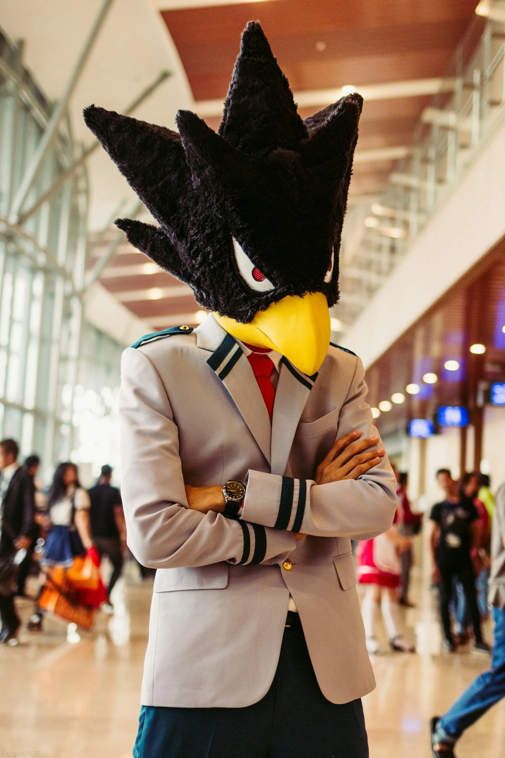 A person wearing a large wolf mascot head with red eyes, dressed in a beige uniform, standing with arms crossed in a busy indoor space.