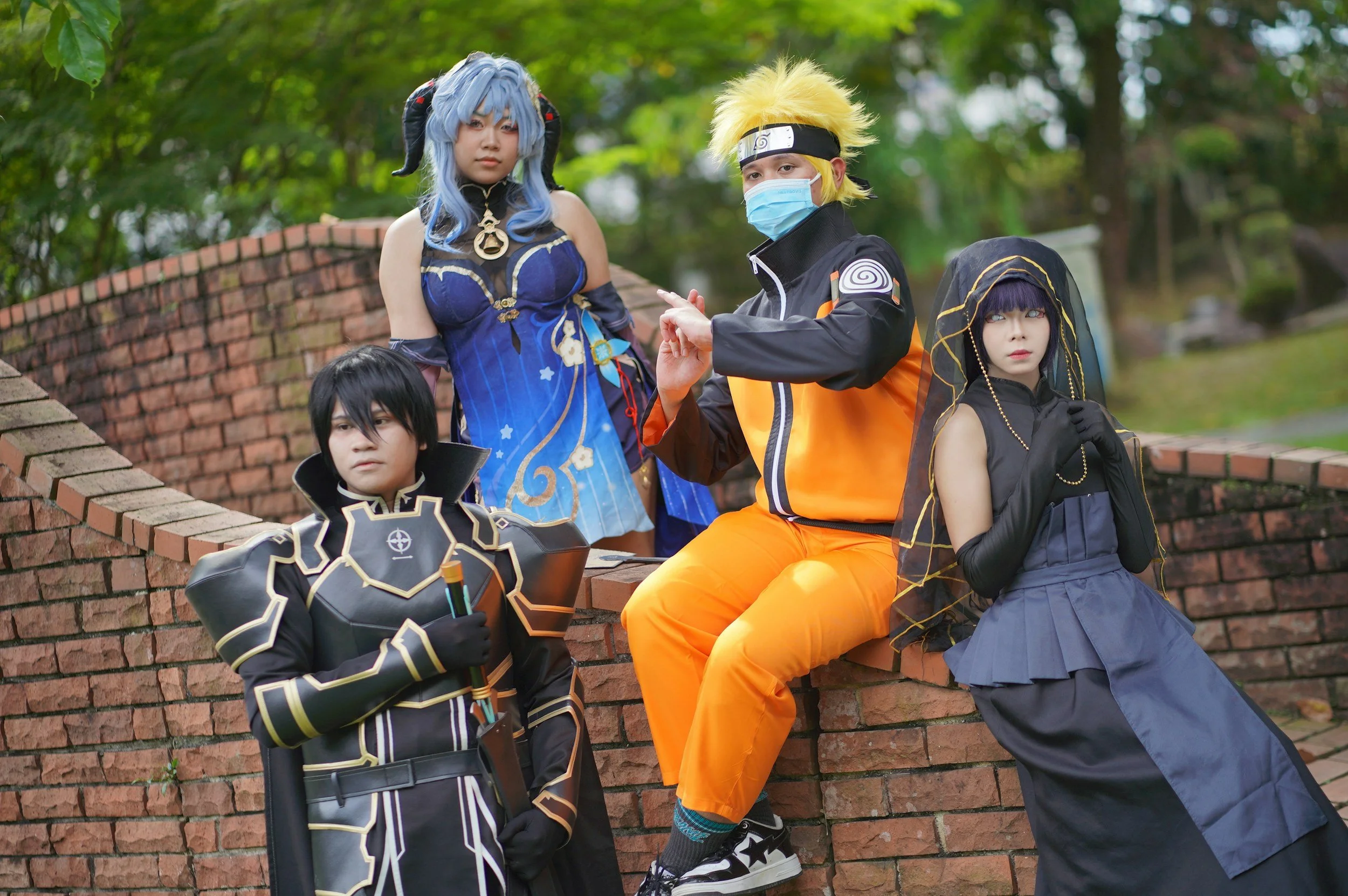 Group of four people in cosplay costumes outdoors near a brick wall, with trees in the background.
