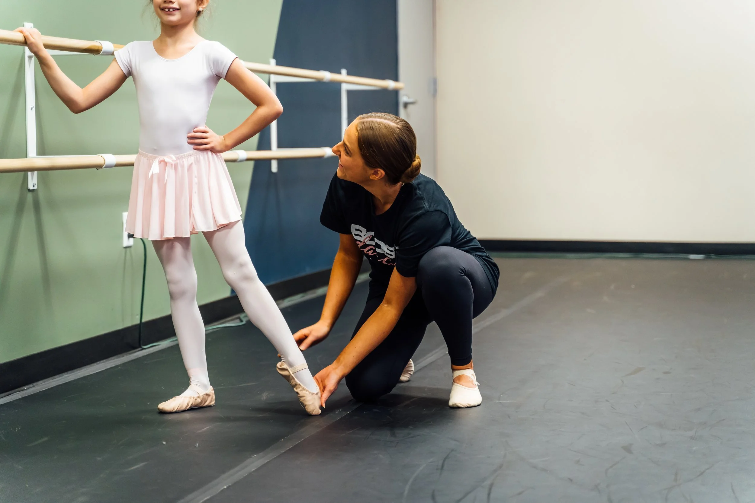 A ballet instructor helping a young girl put on her ballet shoes in a dance studio with a mirror and ballet barres on the wall.