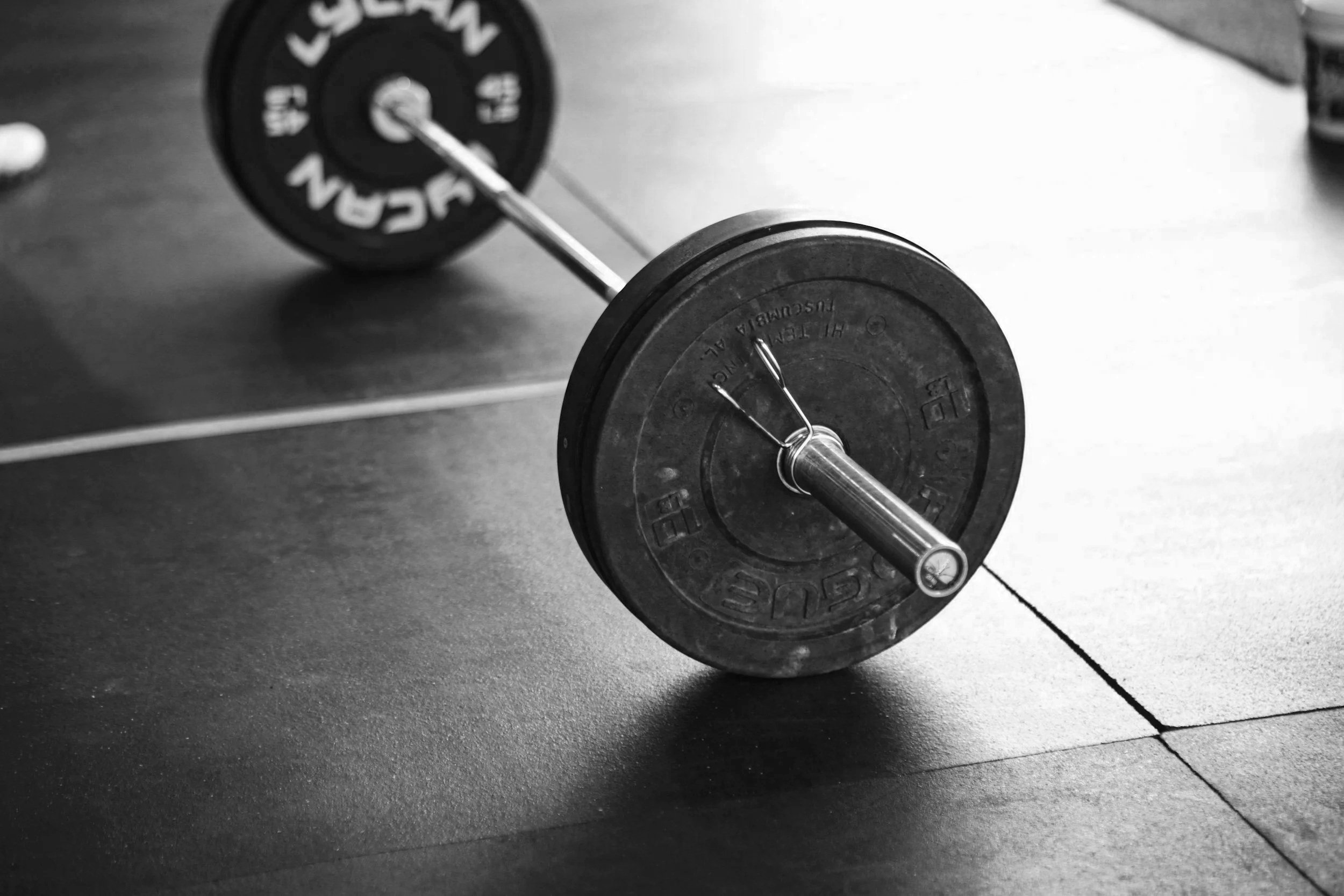 A black and white photo of a barbell with weight plates resting on the floor, with some shadowing and a tiled surface.