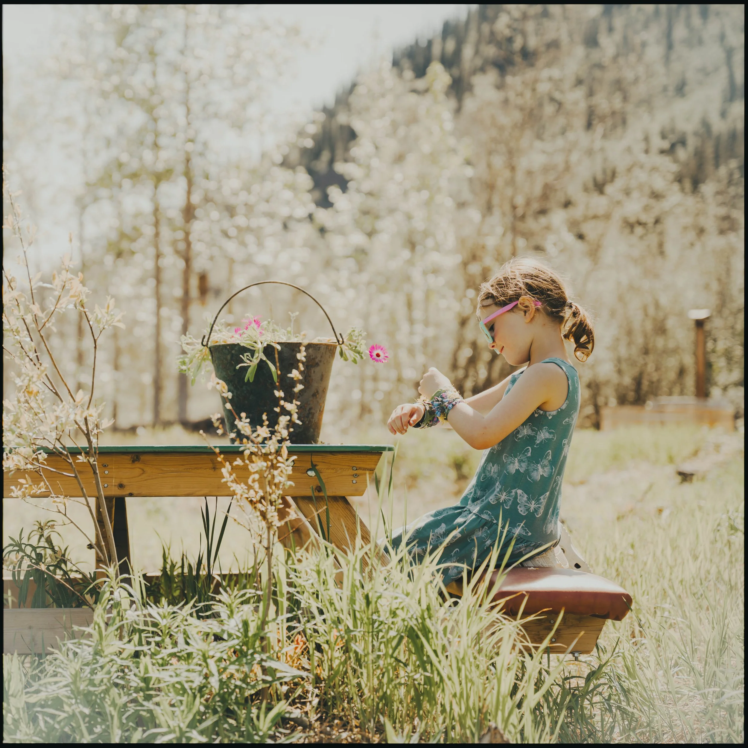 Child sitting at a rustic picnic table in Mt. Lorne, Yukon, making bracelets beside a bucket of wildflowers on a sunny day.