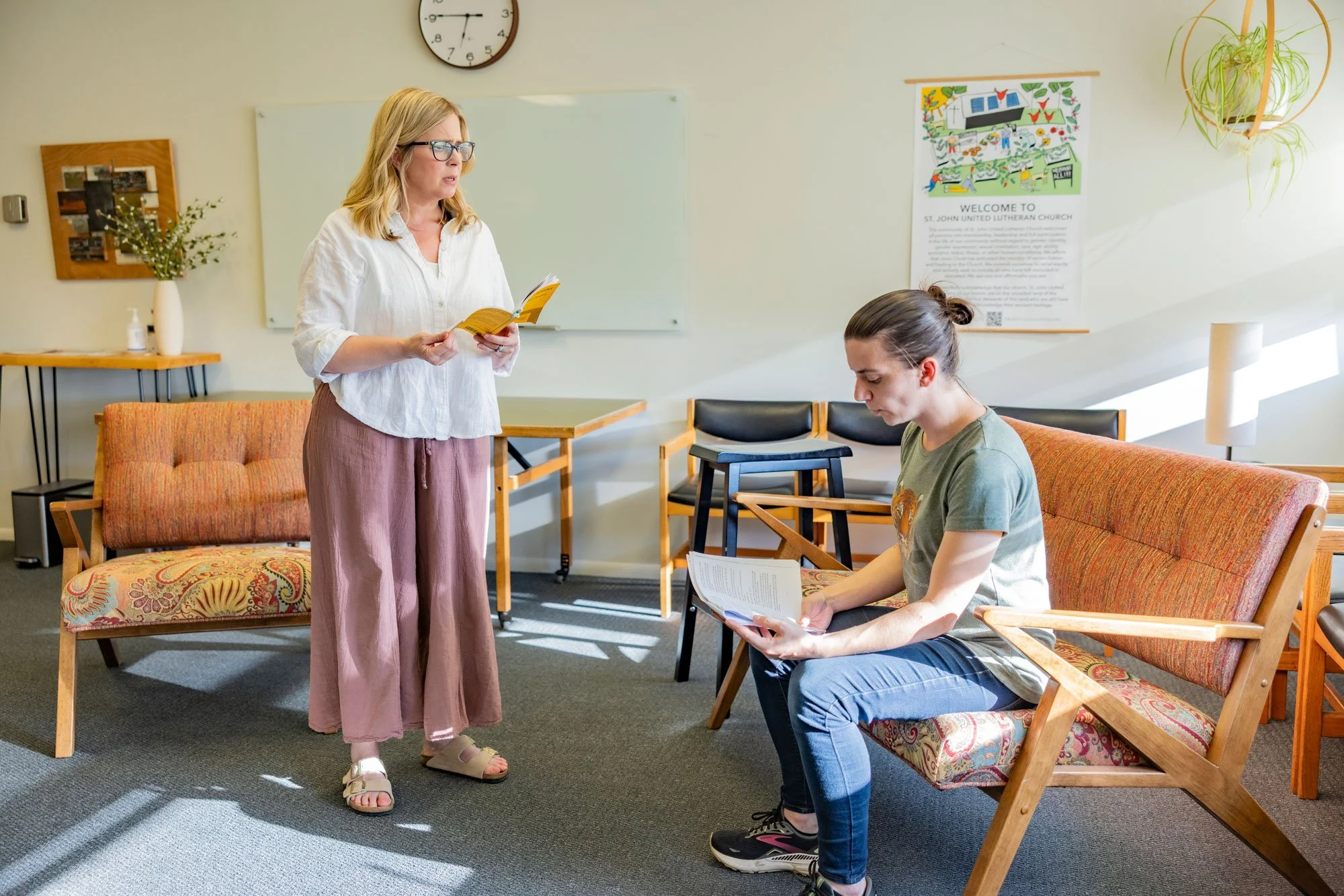 A woman with blonde hair and glasses stands and reads from a play scropt while a young woman with dark hair, tied back, sits on a bench, reading a play script in a room with chairs, a whiteboard, a clock, and a poster on the wall.