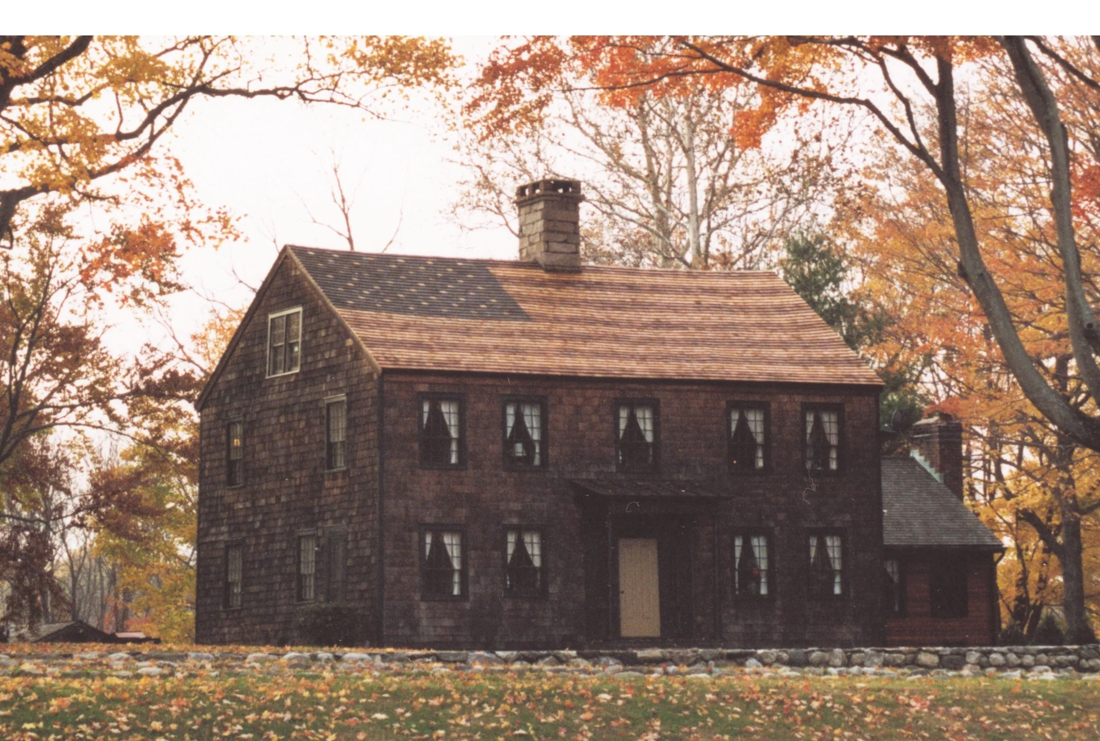 A large, historic brick house surrounded by autumn trees with orange and yellow leaves, with fallen leaves covering the ground.