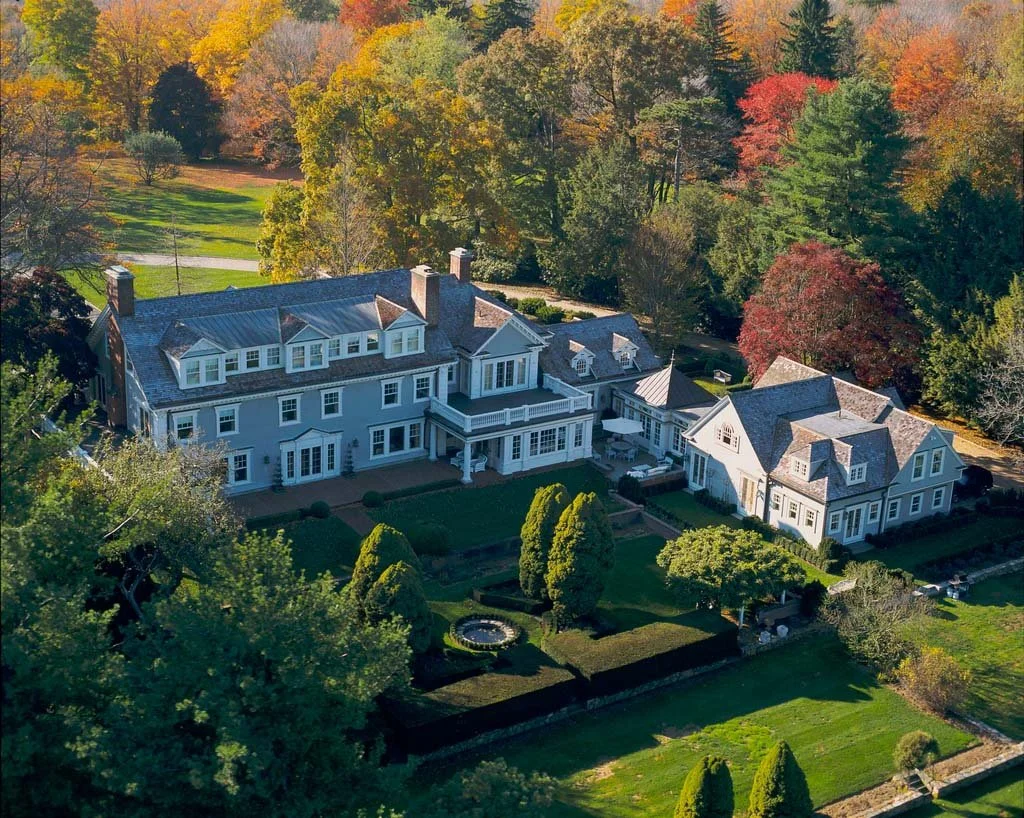 An aerial view of a large, elegant, multi-story house with a well-manicured lawn and gardens, surrounded by colorful autumn trees.