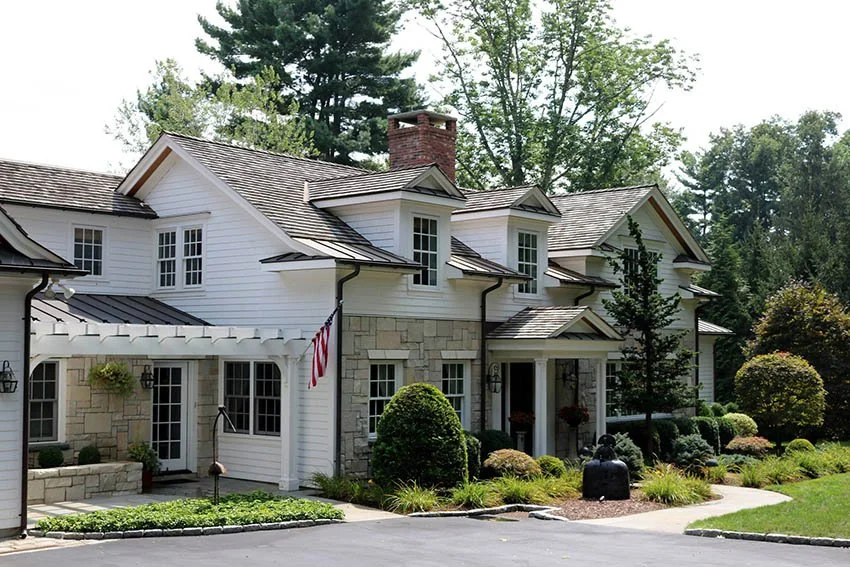 Large white house with multiple gabled roofs, stone and siding exterior, surrounded by landscaped garden with bushes, trees, and a driveway.