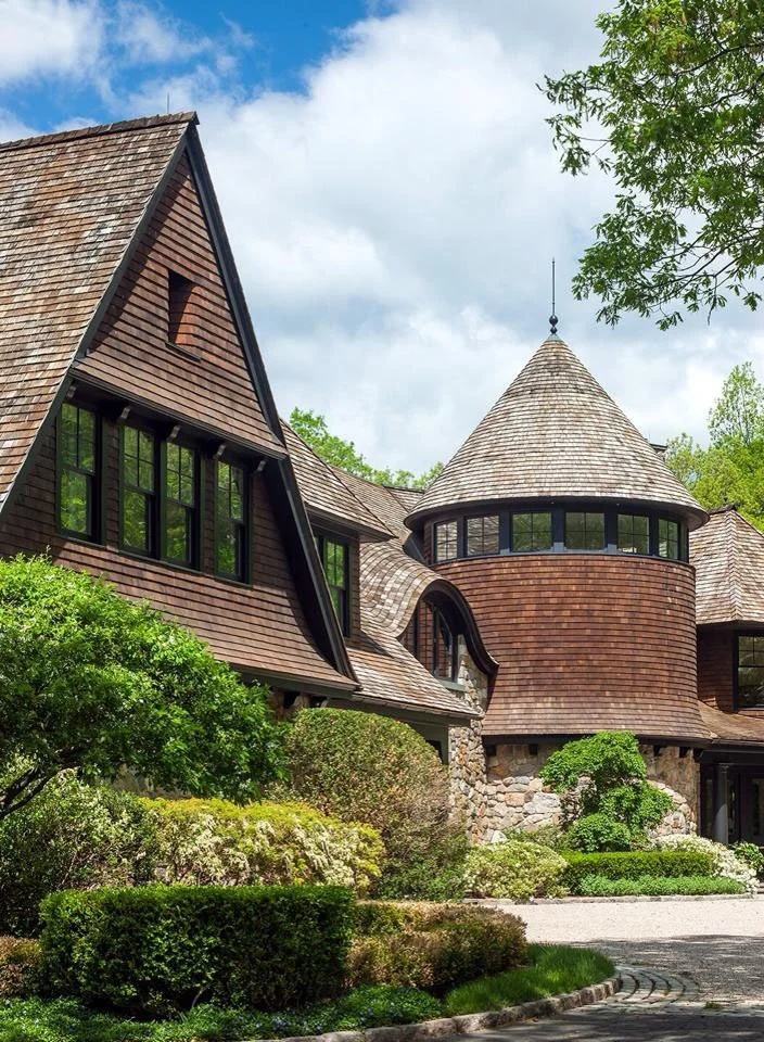 A large house with a steeply pitched and curved roof, multiple windows, and surrounded by lush green landscaping.