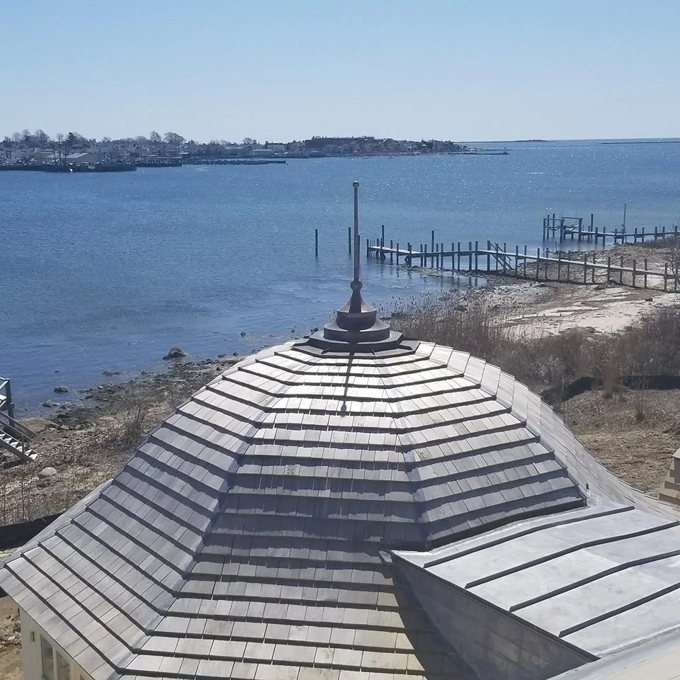 View of a roof with a metal flagpole ornament, overlooking a calm body of water with docks and a distant shoreline on a clear day.