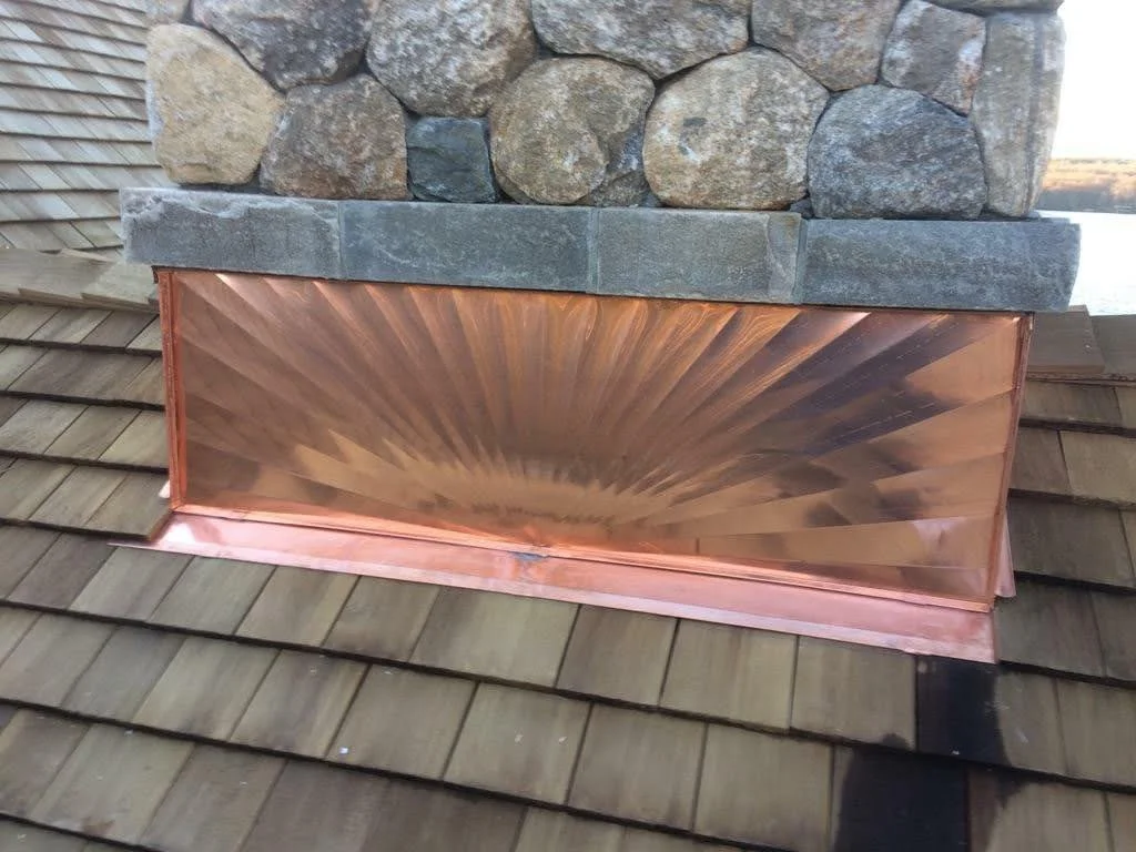 Copper chimney cap with sunburst pattern on a shingled roof, in front of a stone chimney.