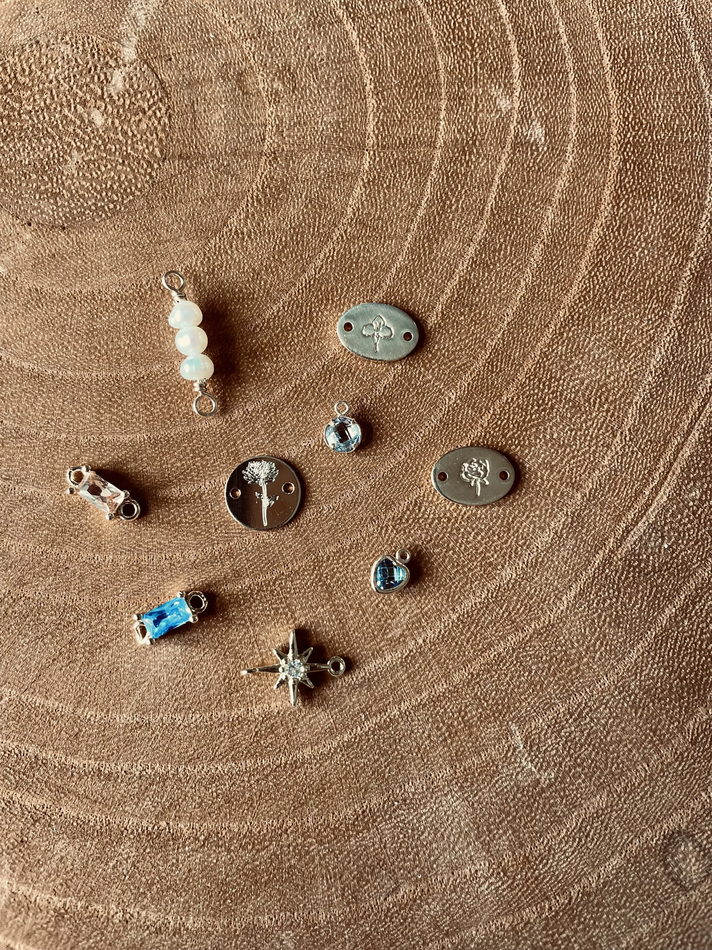 Various small decorative jewelry charms and pendants on a wooden surface, including pearls, a starburst with a crystal, heart, and oval-shaped pieces with floral designs.