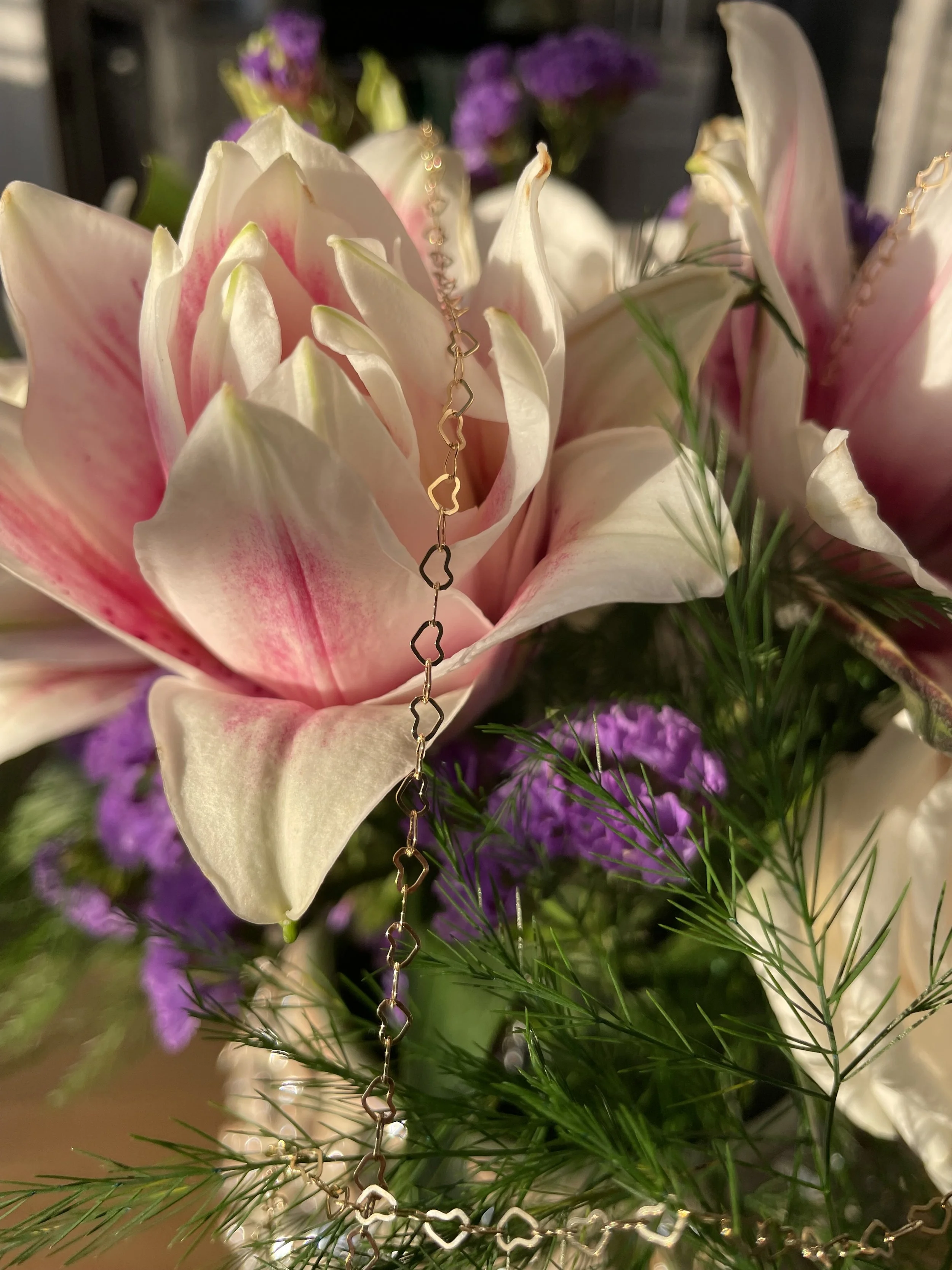Close-up of a floral arrangement with a pink and white flower, purple flowers, and green foliage, with a gold chain draped over the pink and white flower.