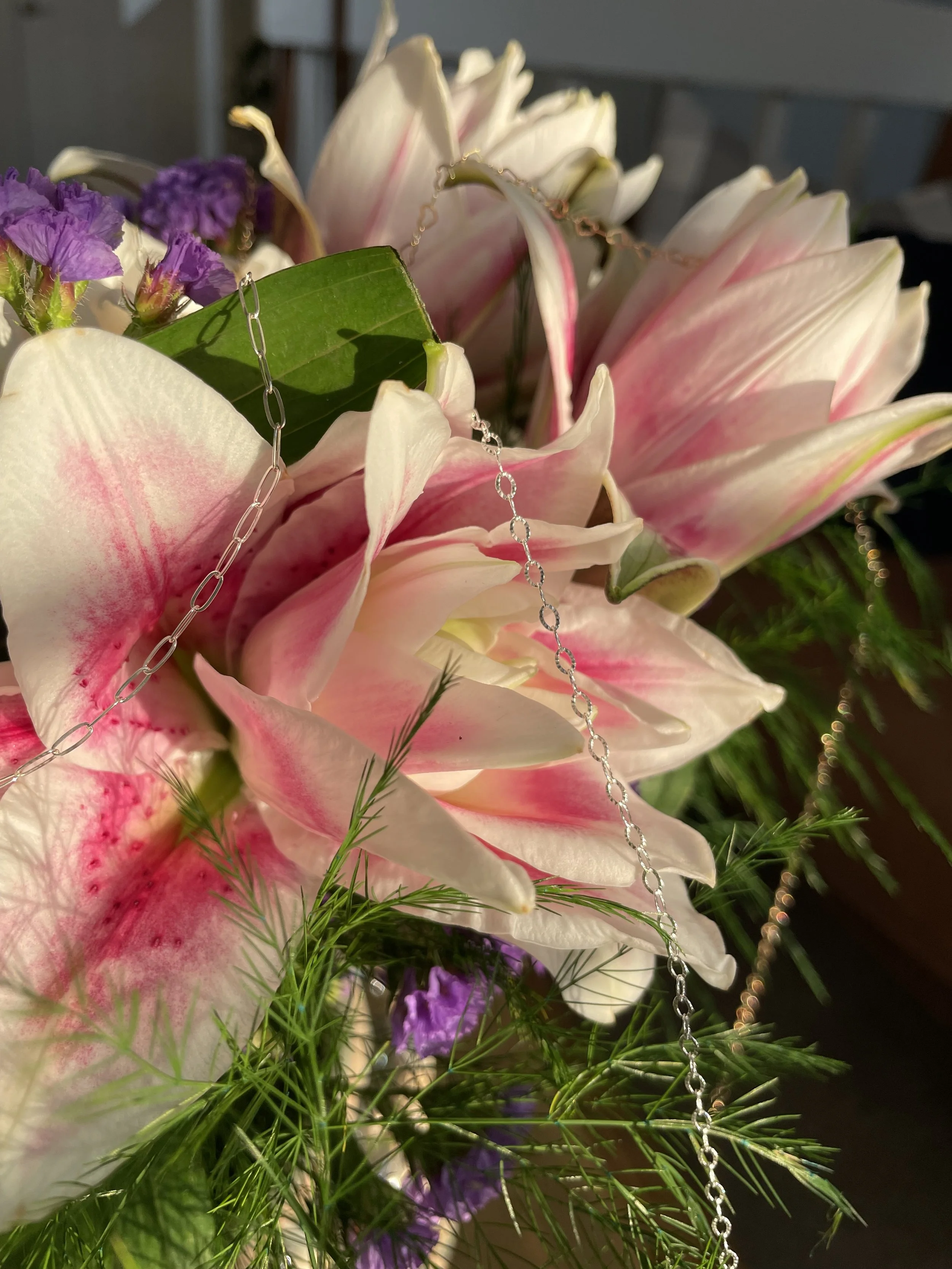 Close-up of a flower bouquet with pink and white lilies, small purple flowers, green leaves, and delicate silver chains.