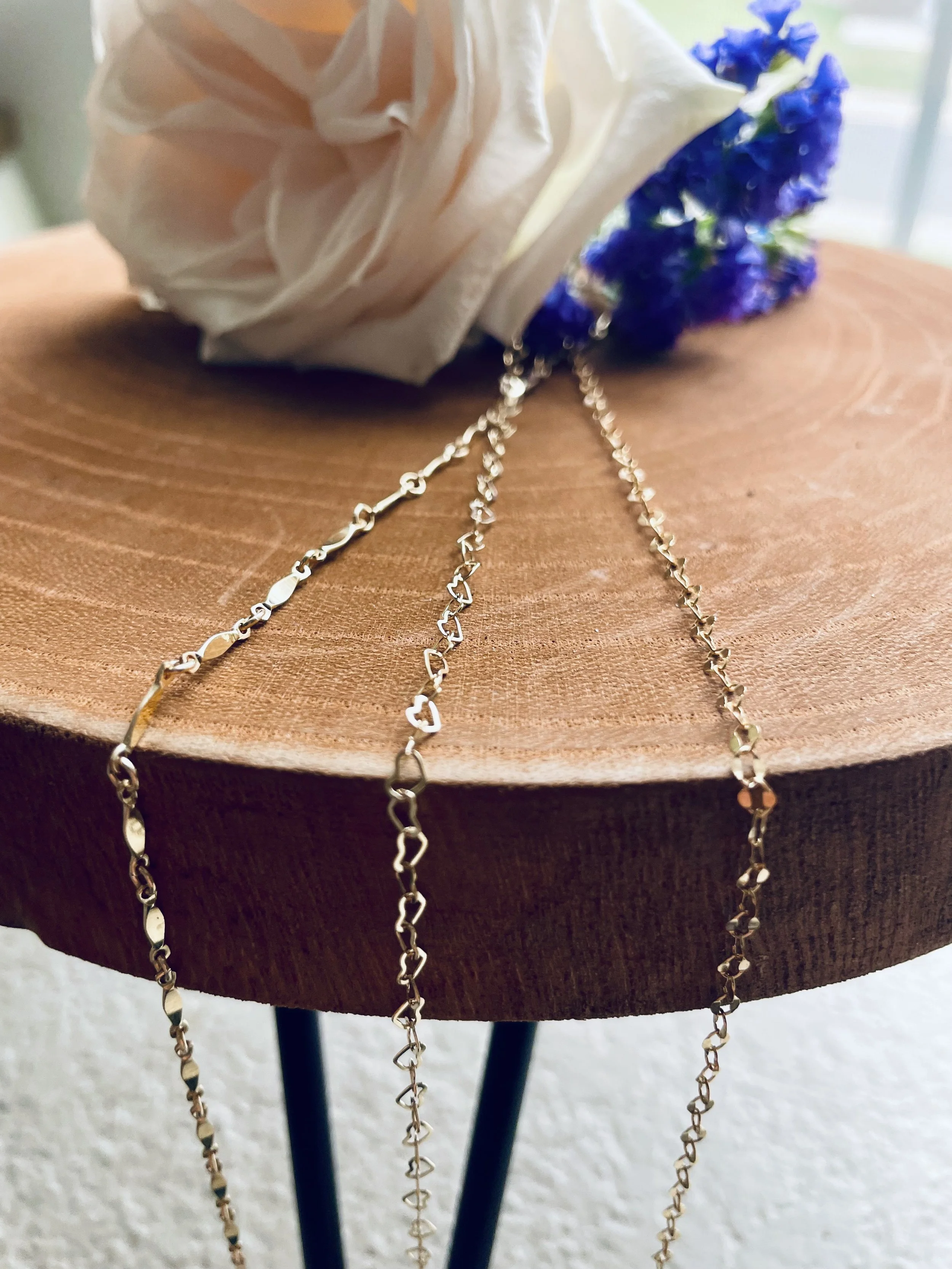 Three delicate silver necklaces with intertwined heart and oval links, draped on a round wooden table. A white flower and a blue flower are placed beside the necklaces.