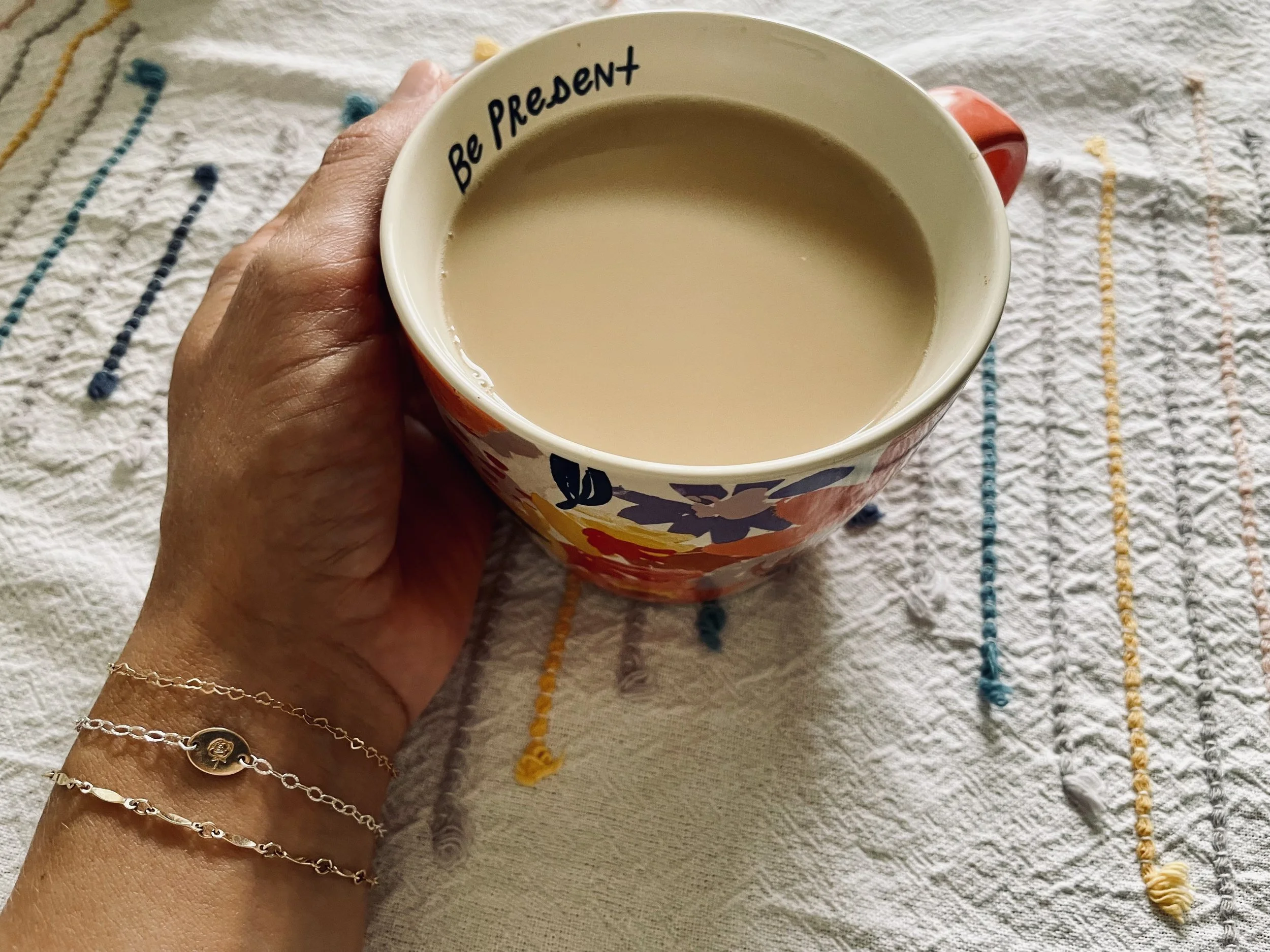 A hand holding a colorful mug filled with a light brown beverage on a textured, embroidered white cloth with multicolored thread designs, and the mug has the phrase "Be Present" written on the inside rim.